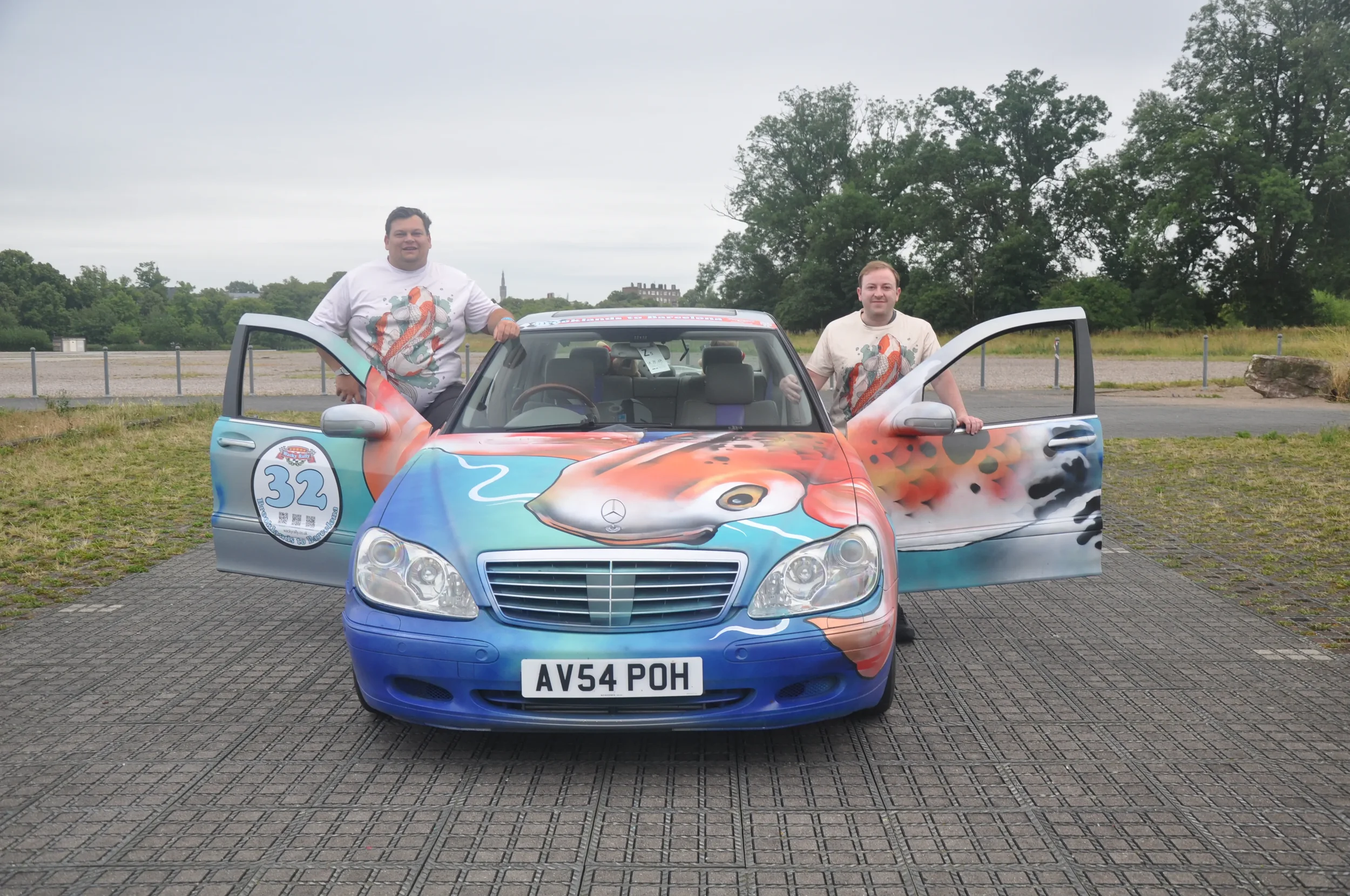 Two men standing next to a Mercedes-Benz car with a colorful, airbrushed race car theme. The car has a picture of a fish on the hood, a racing number 32 on the side, and artistic flames on the doors. The men are holding the open car doors, standing o
