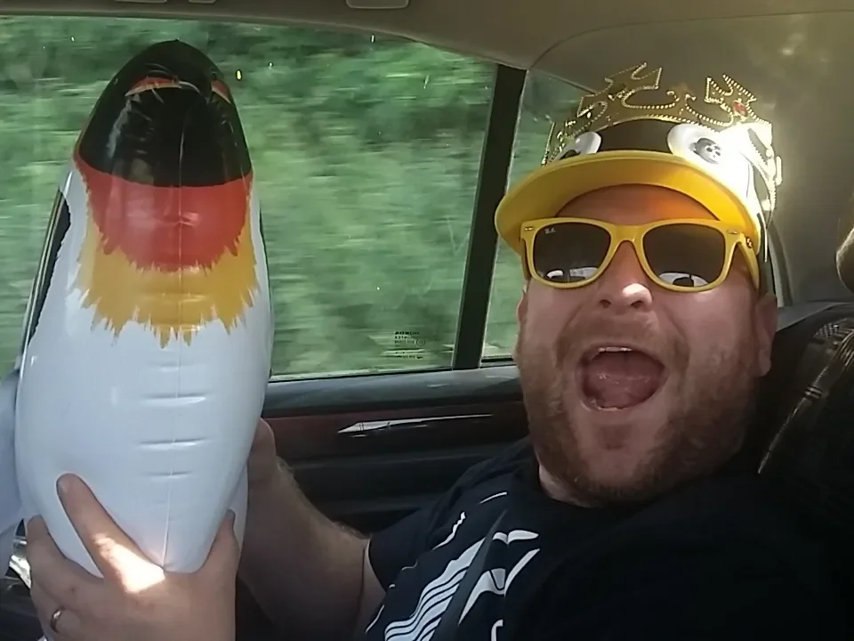 A man wearing yellow sunglasses and a crown-shaped headband sitting in a car, enthusiastically holding and waving a large inflatable penguin.