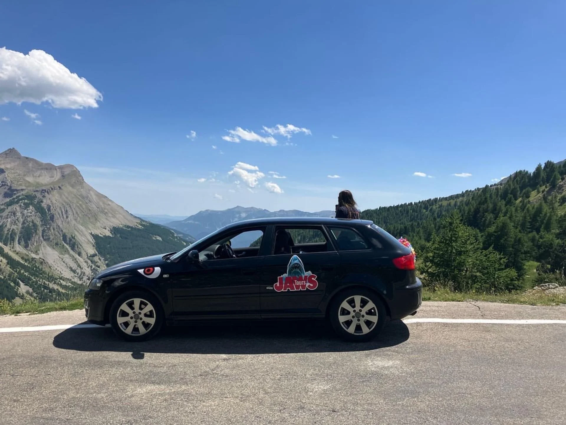 A black car with a JAWS-themed sticker parked on a mountain road overlooking a scenic mountain landscape with green trees and blue sky.