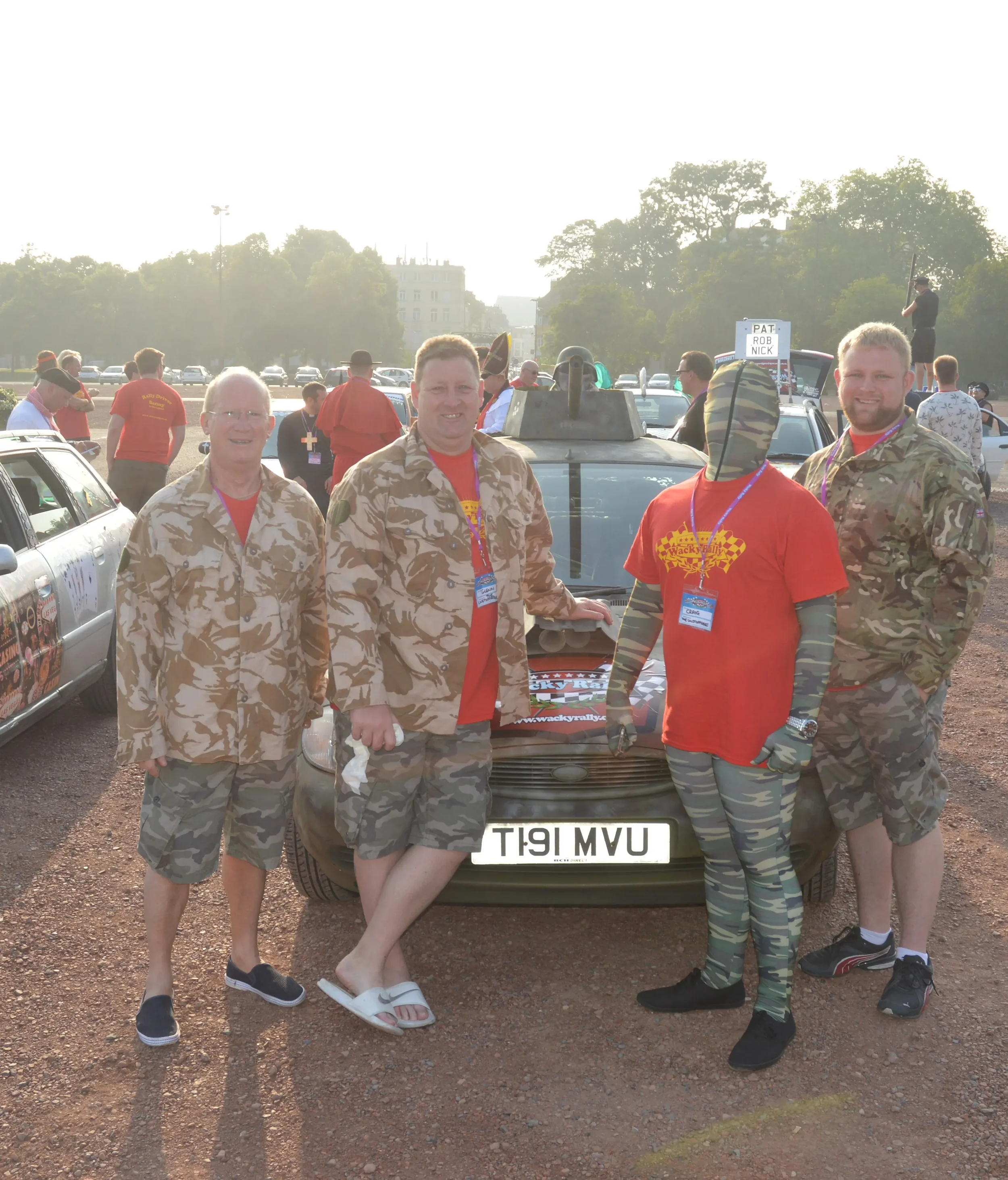 Group of five men posing in front of a military-style car at an outdoor event, some dressed in camouflage military jackets and others in casual red t-shirts, with cars and people in the background.