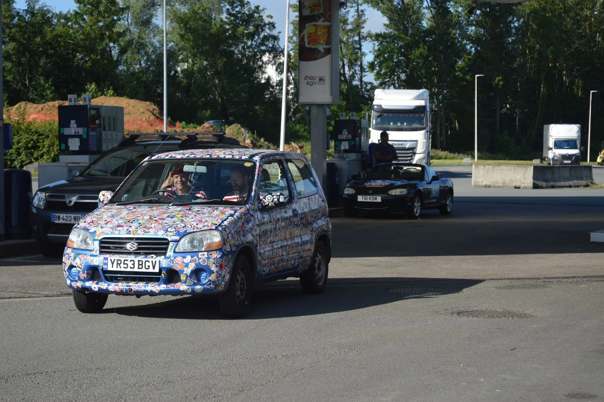 A colorful, sticker-covered Suzuki hatchback at a gas station with a person in the driver's seat, two trucks, and another small car in the background.
