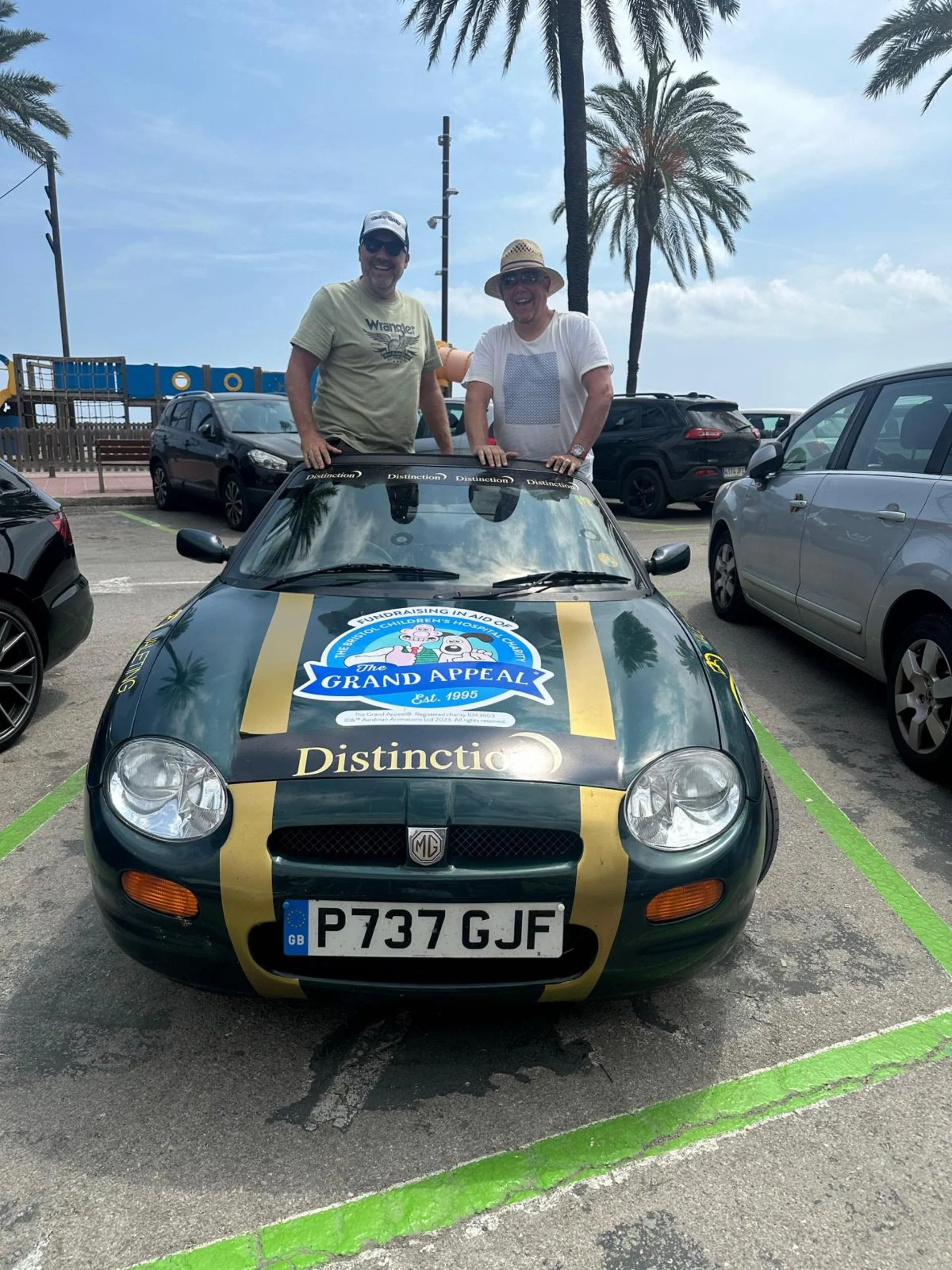 Two men standing in a convertible car with a racing style paint job, parked in a parking lot with palm trees in the background, on a partly cloudy day.