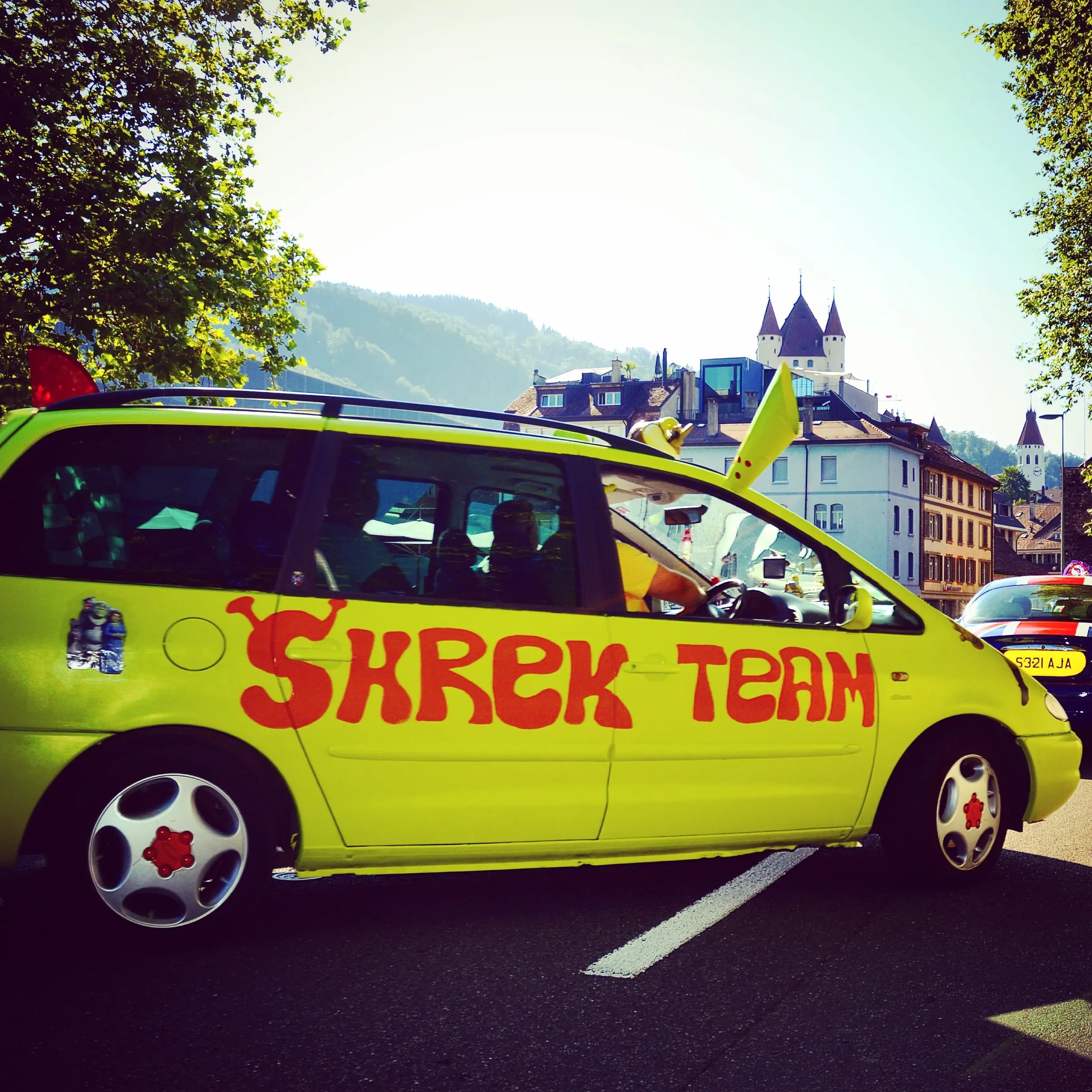 A bright yellow car with red lettering that says 'SHREK TEAM' parked in a lot with other vehicles. The car has novelty hubcaps and is decorated with Shrek character stickers. In the background are trees, a historic town with buildings, and a castle o