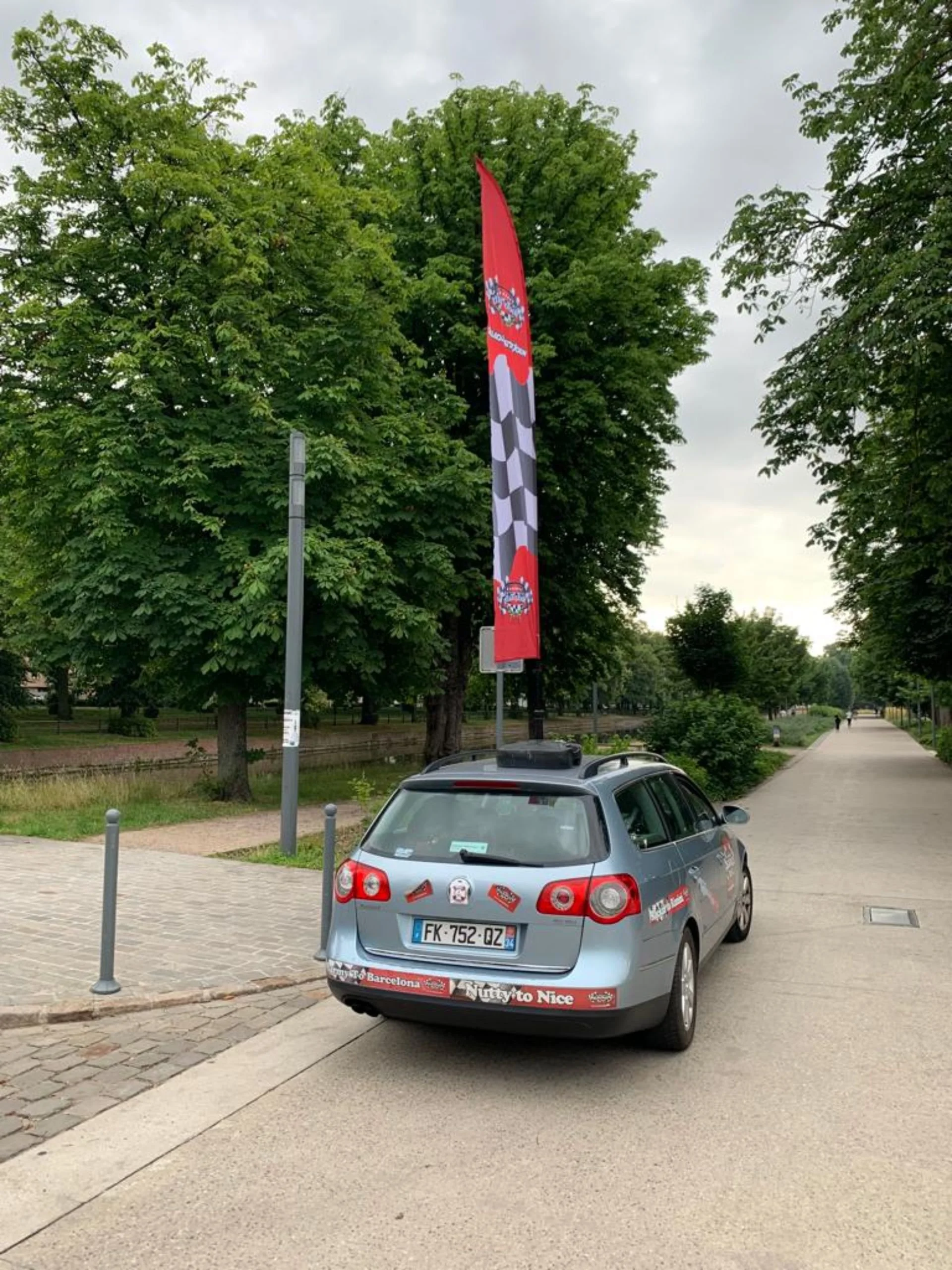 A silver station wagon parked on the side of a wide sidewalk with a tall red and black flag attached, decorated with racing checkered patterns and various stickers, including promotional ones.