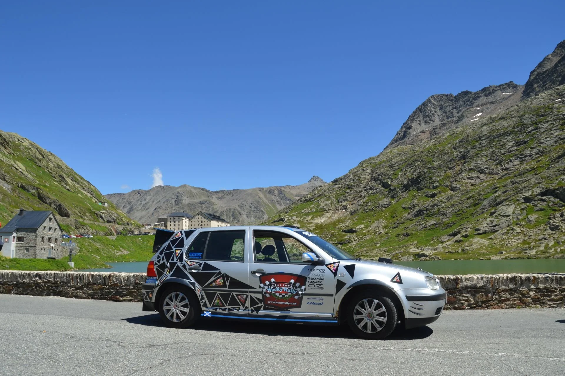 A painted rally car parked on a mountain road with rocky green hills, a lake, and buildings in the background under a clear blue sky.