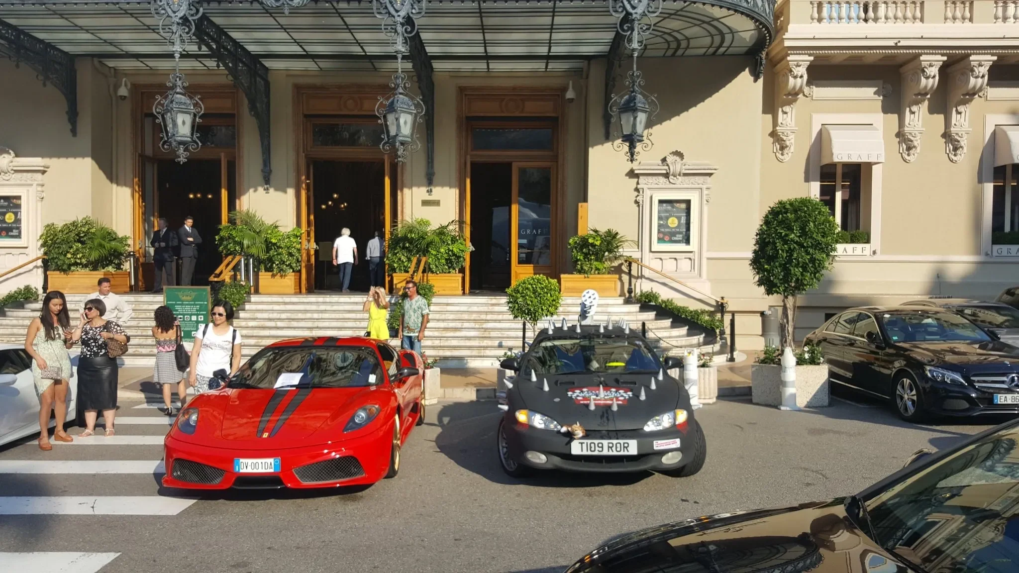 People gathering outside a building with classic architecture, three luxury cars parked in front, including a red sports car and a black car with decorative spikes, while others are walking on the sidewalk and stairs.