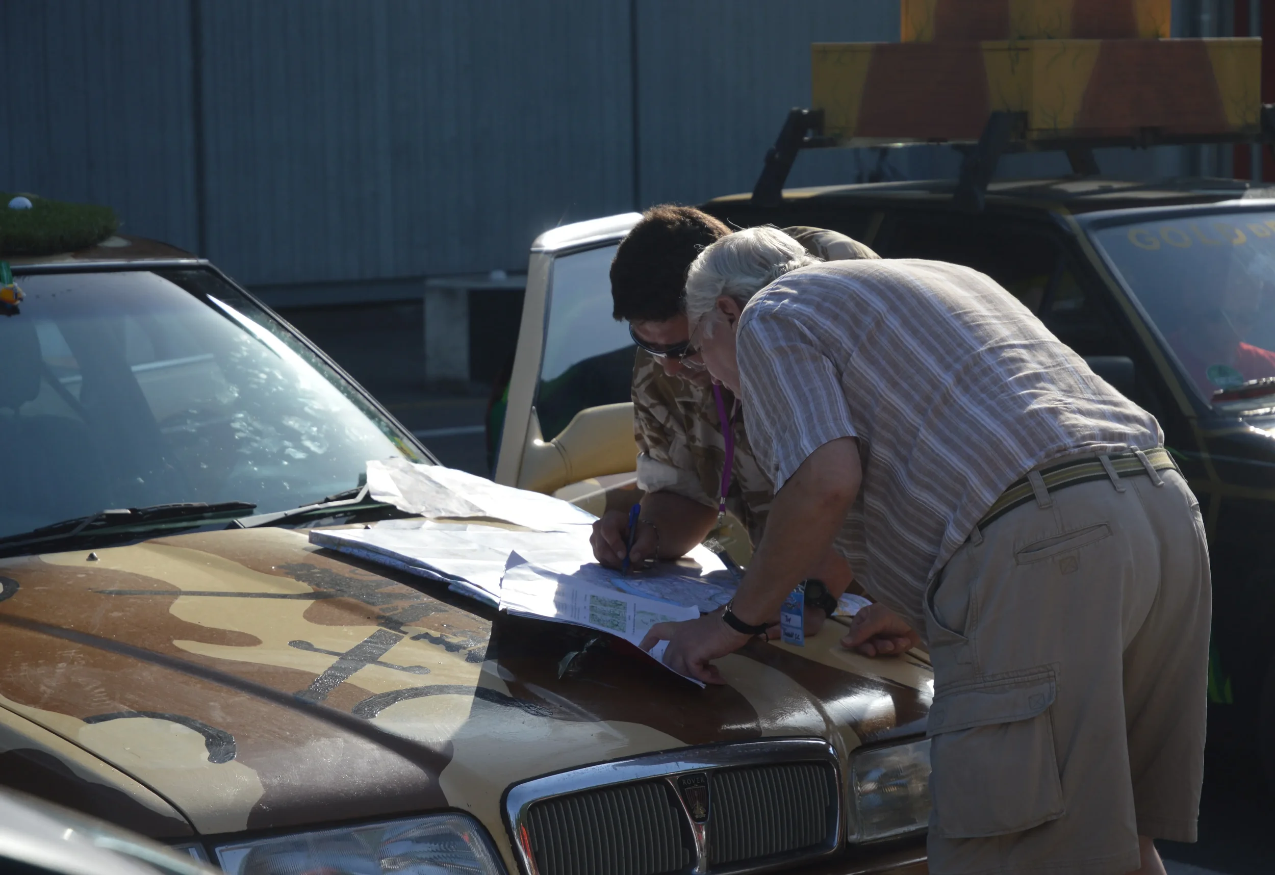 Two men examining documents on the hood of a camouflage-colored emergency vehicle, with other vehicles and a fence in the background.