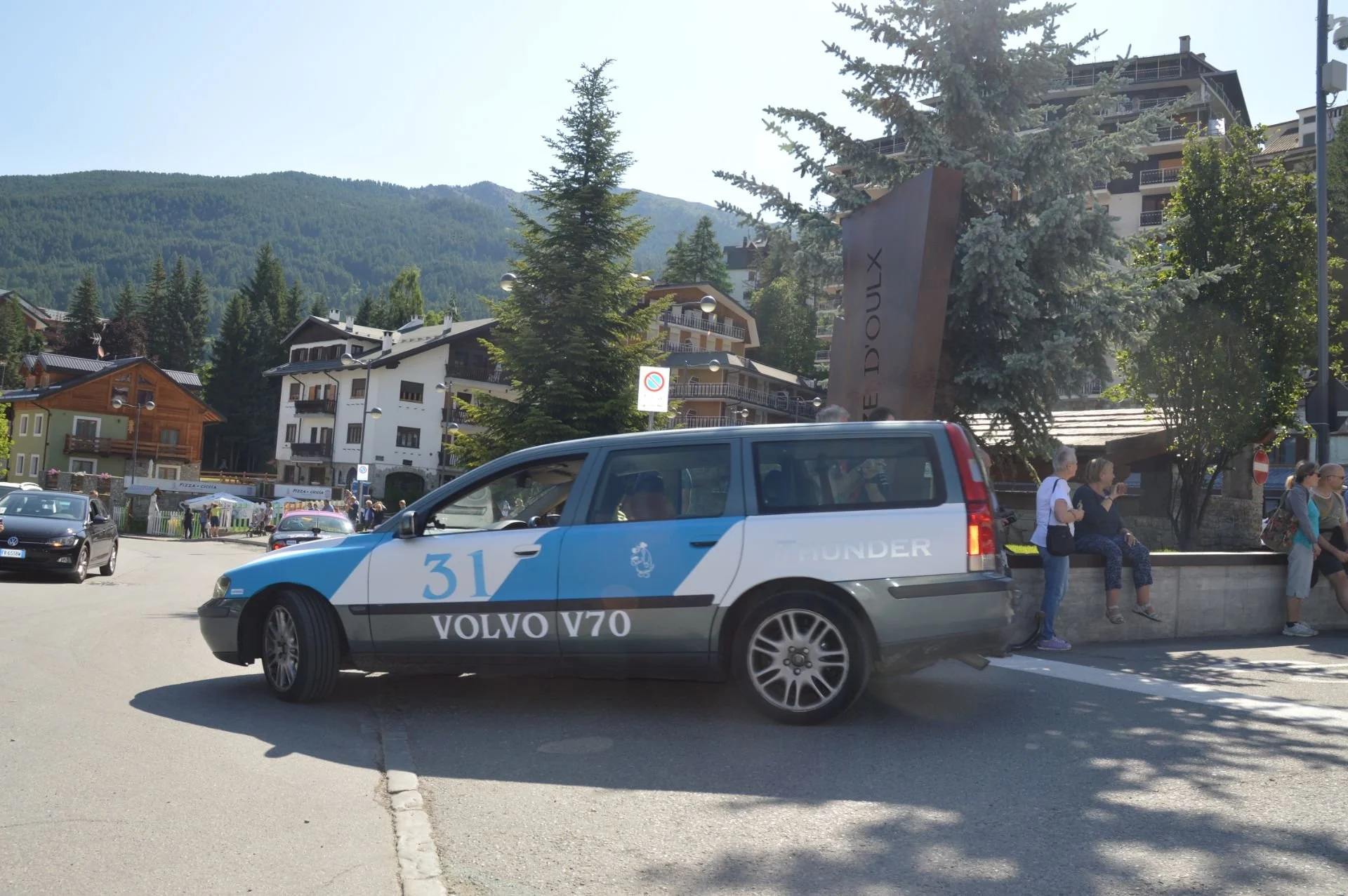 A gray and blue police car with 'VOLVO V70' written on the side, parked on a street in front of a mountain town with people sitting on a ledge nearby and residential buildings in the background.