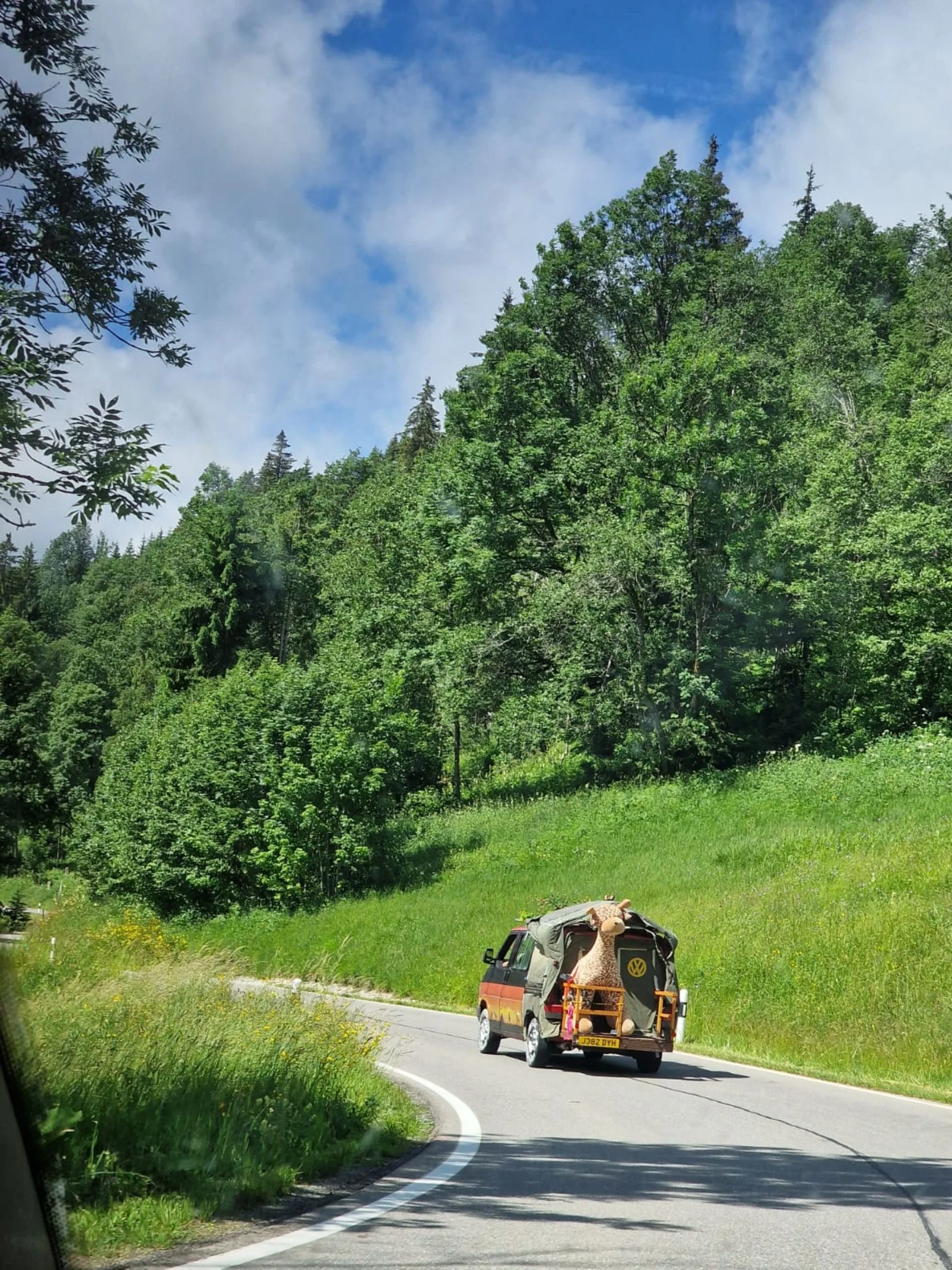 A van traveling on a curved mountain road surrounded by lush green trees and grass, with a large giraffe mascot figure attached to the back of the van.