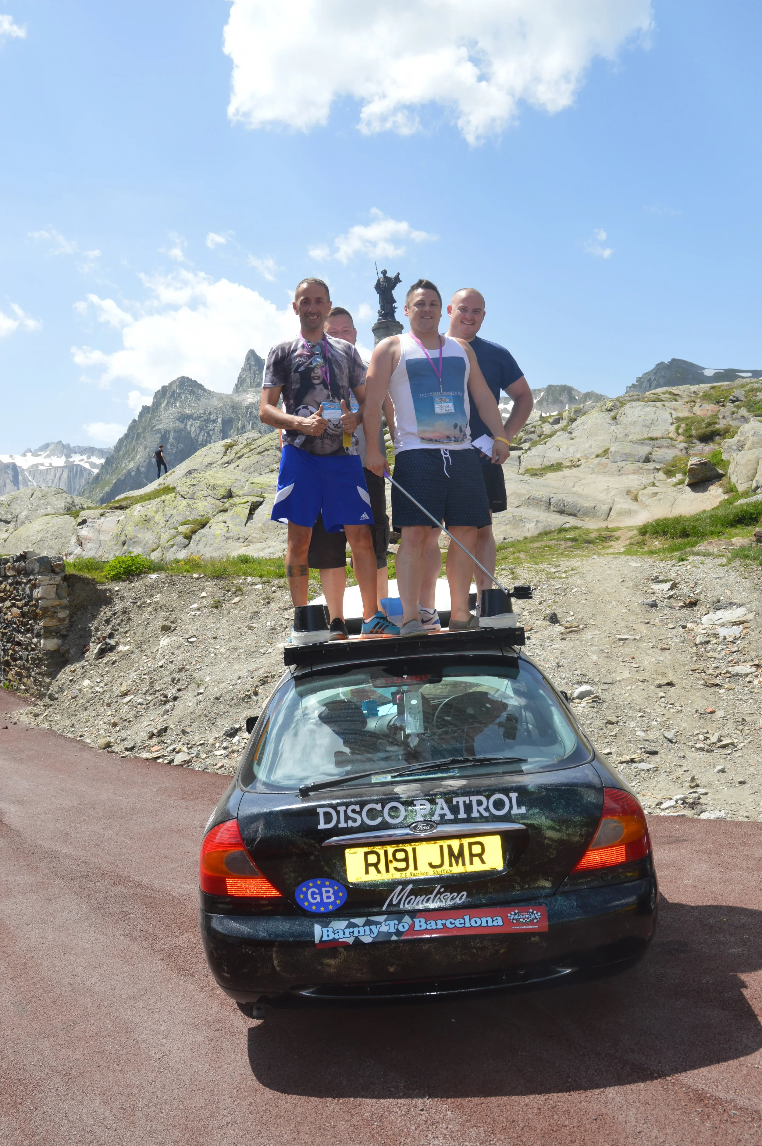 Four men standing on top of a black car labeled 'Disco Patrol,' with a scenic mountainous background and a statue of a figure on horseback on a hill in the distance.