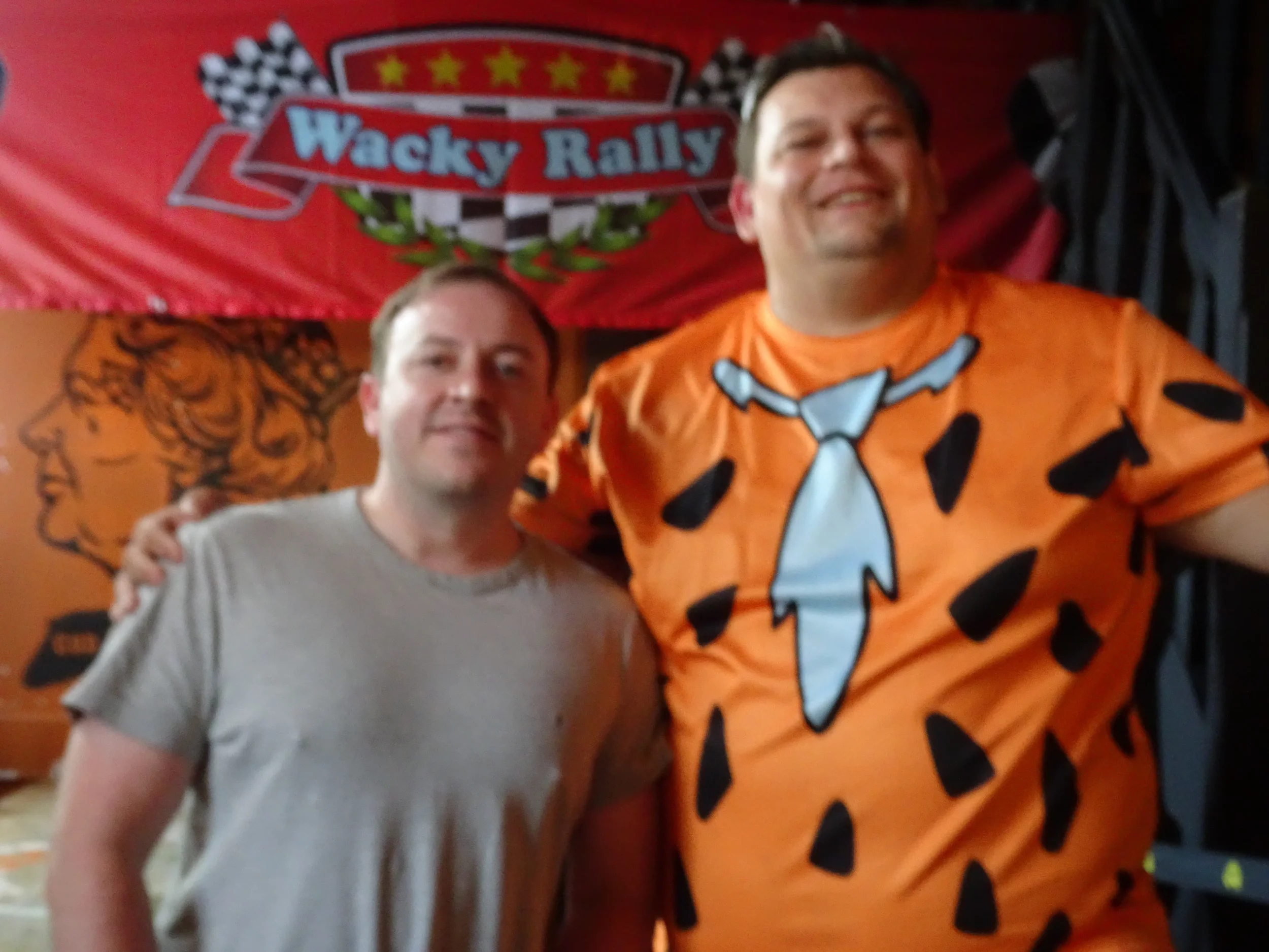 Two men posing together in front of a red banner with a racing logo that says Wacky Rally. One man is wearing a beige t-shirt, and the other is wearing an orange t-shirt with a cartoonish bald head and suit tie design.