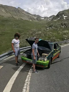 Two boys standing by a green and black car with its hood open, on a mountain road with a scenic mountainous backdrop.