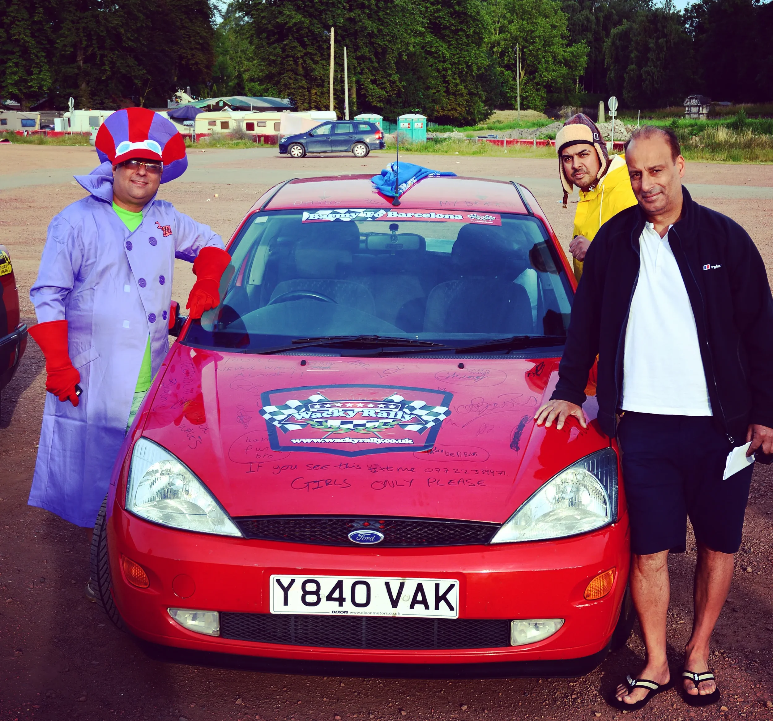 Three men standing next to a red car at a rally event, one dressed in a colorful clown costume with a large hat, another in a yellow jacket with a helmet, and the third in a black jacket and shorts, with signatures and messages written on the car's h
