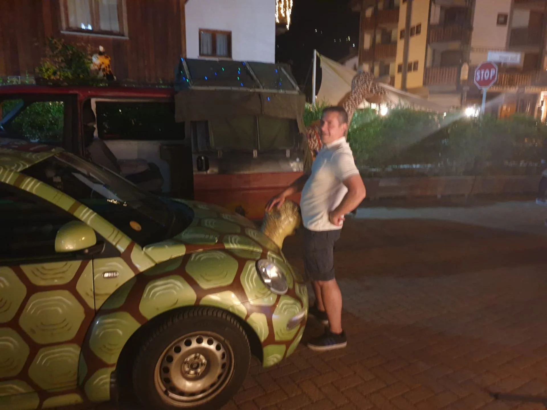 A man standing next to a car with a turtle shell design, on a nighttime city street.