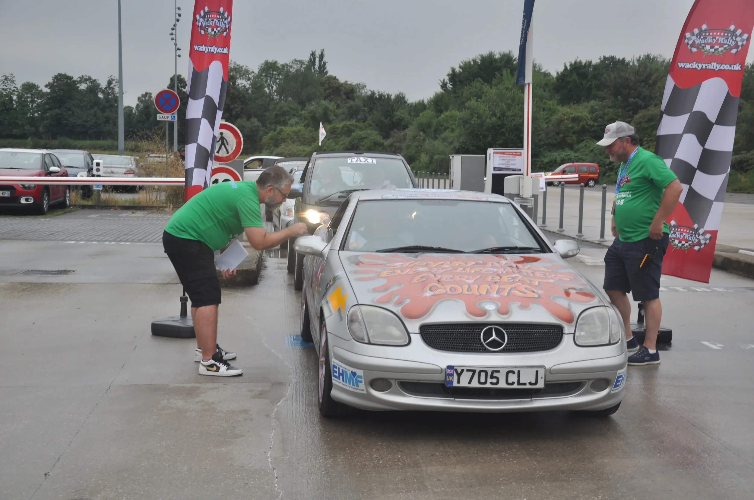 Two men in green shirts cleaning a silver Mercedes-Benz race car with a painted Hello Kitty design on the hood at a rally event, with flags and other vehicles in the background.