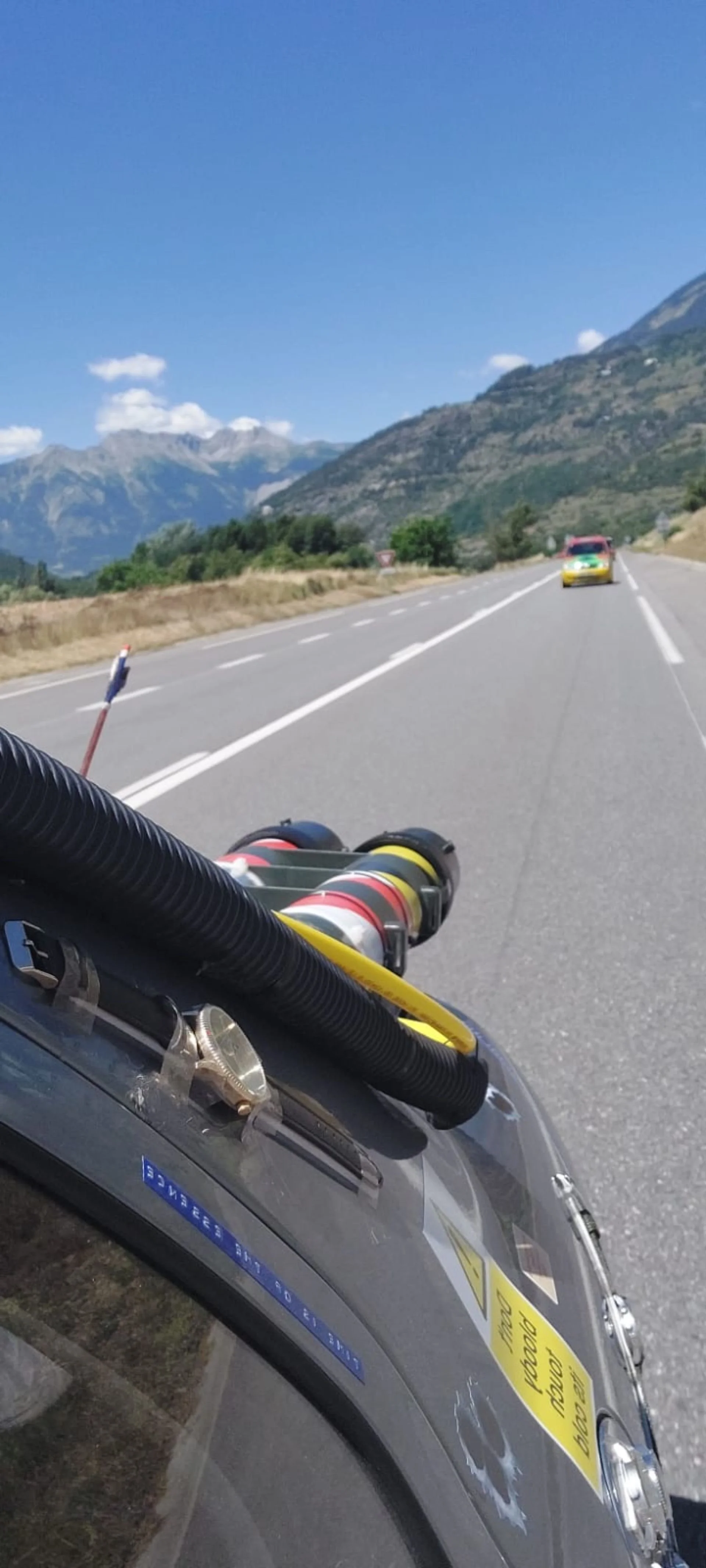 Mountains in the background with a clear blue sky, a yellow race car on the road ahead, and part of a silver race car with safety equipment and stickers in the foreground.