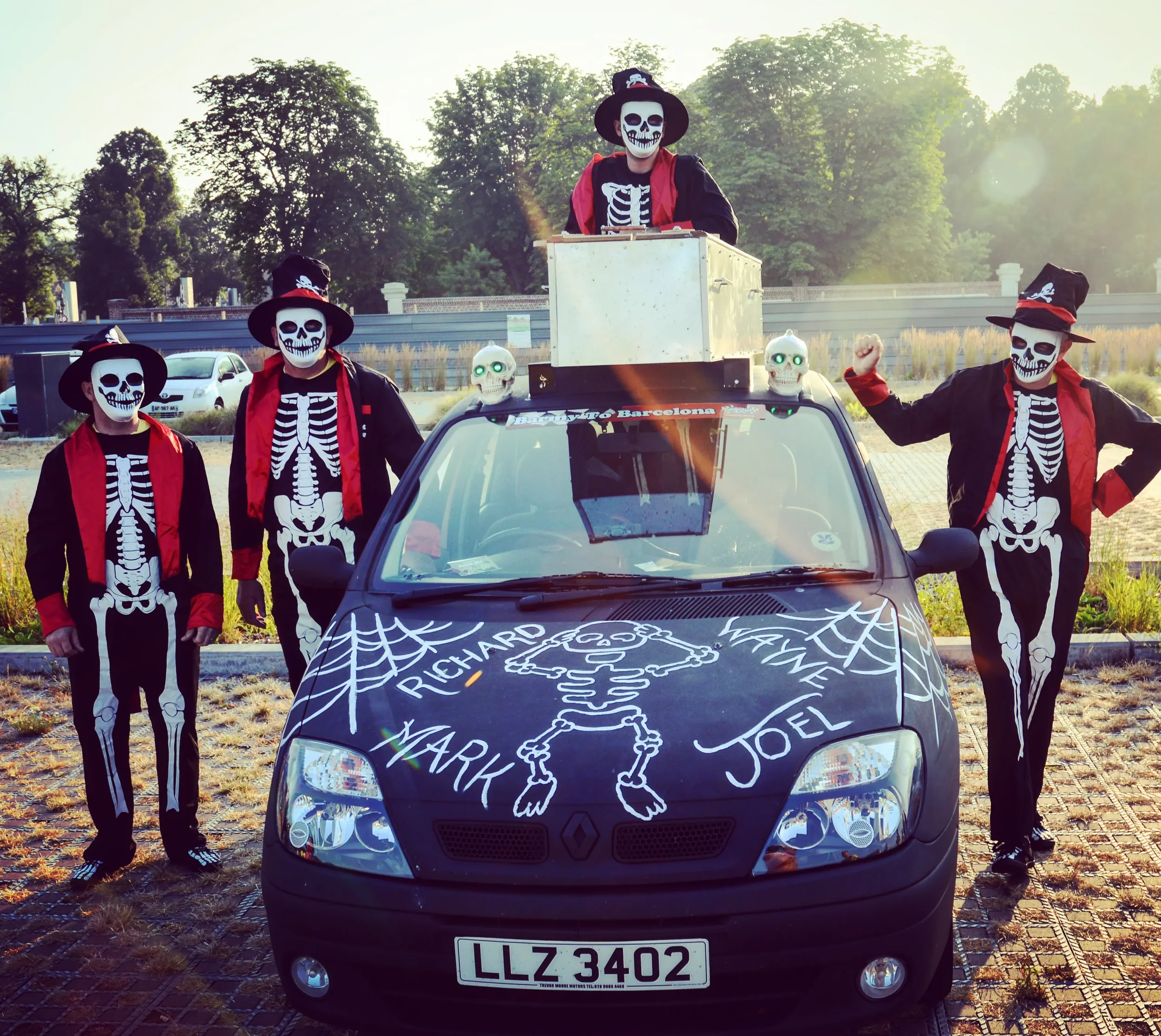 Five people dressed in skeleton costumes with masks and hats, standing around a car decorated for Halloween. Two are on the ground, one is standing on the driver's side, one is standing on top of the car, and one is on the passenger side with one arm
