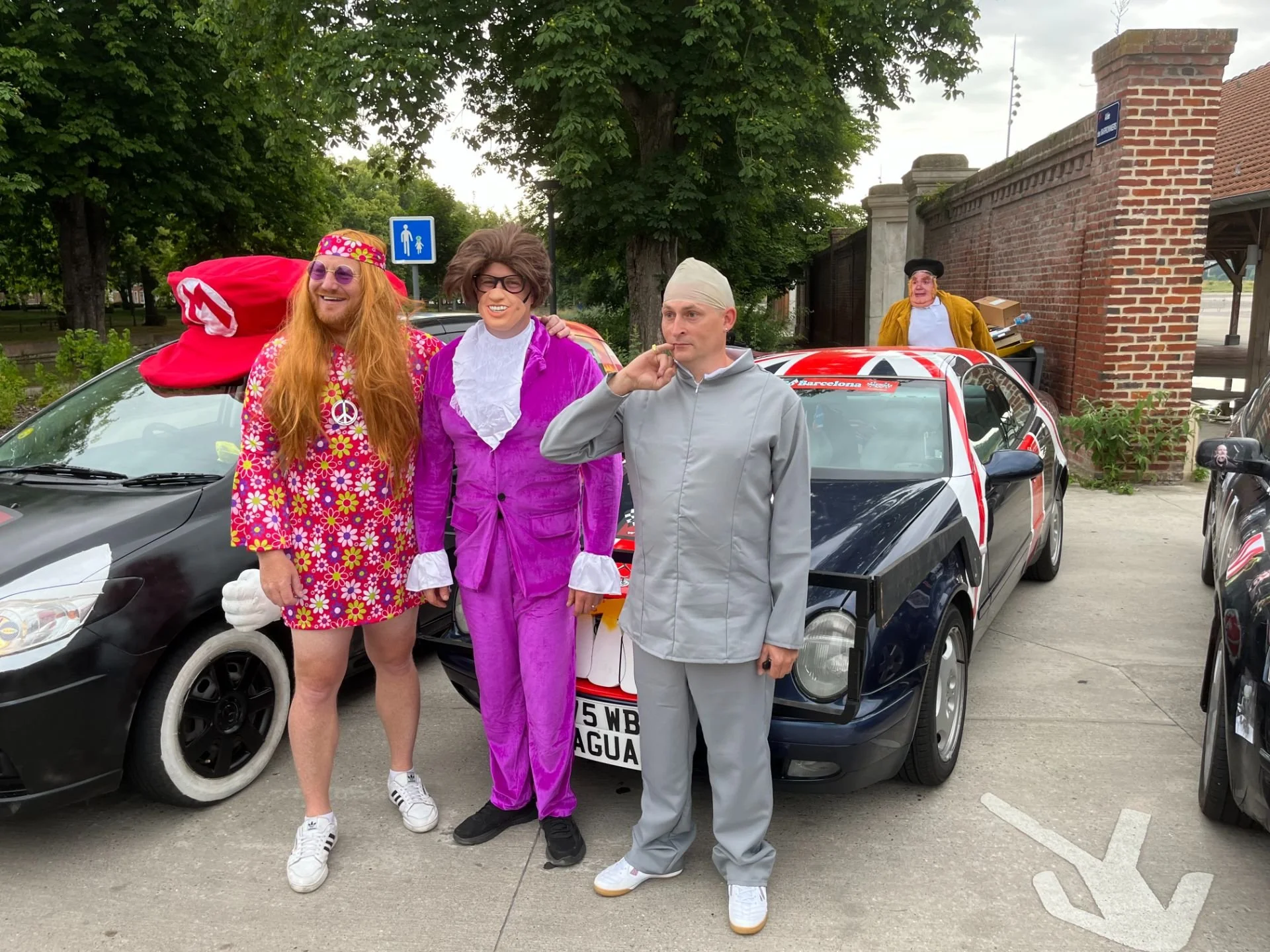 Four people dressed in colorful, eccentric costumes standing in front of a black car with a Union Jack flag design. One person is wearing a pink floral dress and hat, another in a purple suit with glasses, a third in a gray tracksuit with a bandaged 