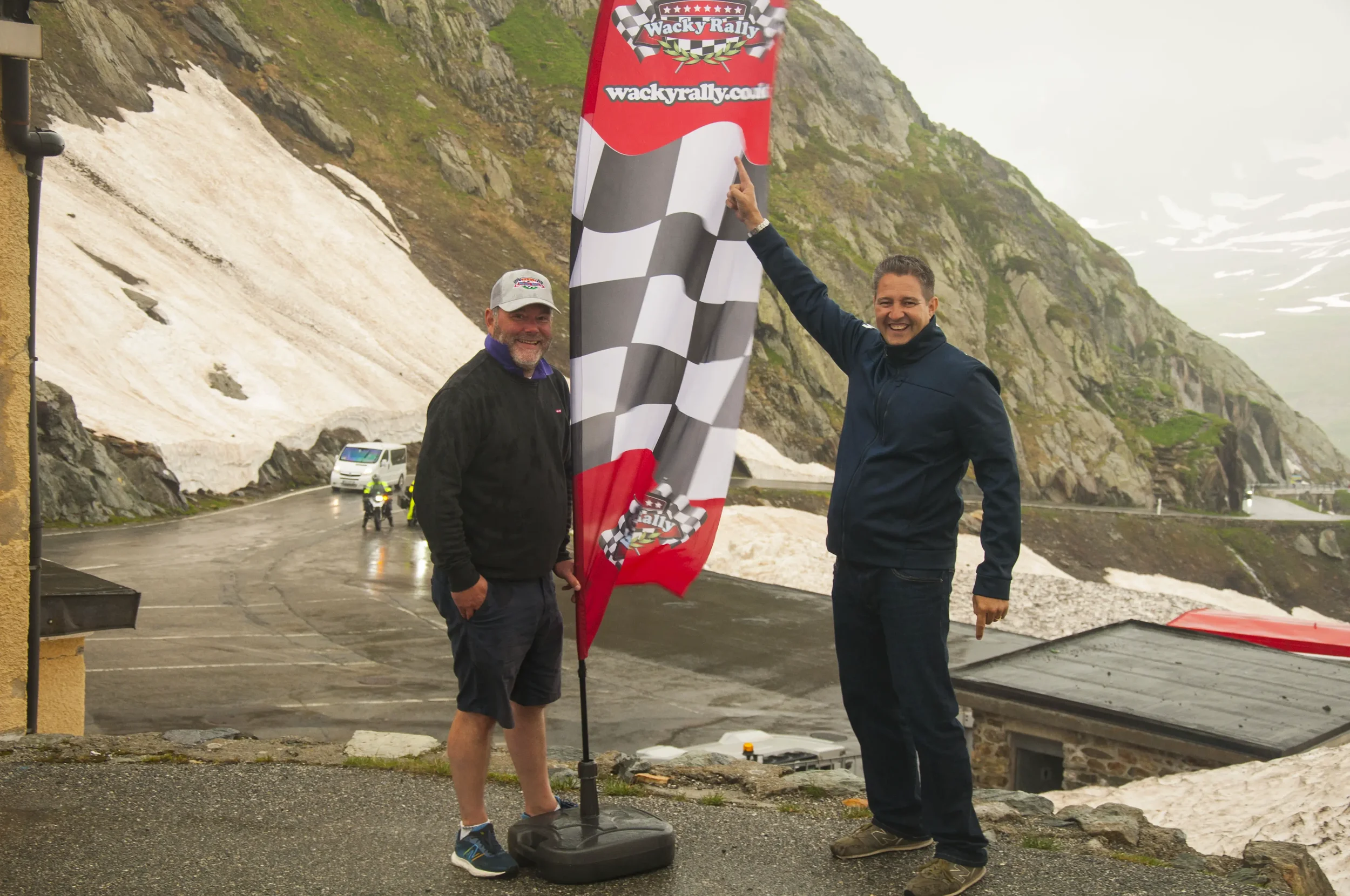 Two smiling men standing outdoors next to a checkered flag banner. The man on the right is pointing at the flag, and the man on the left is wearing a cap and shorts. The background shows a mountain with patches of snow and a wet road, indicating cool