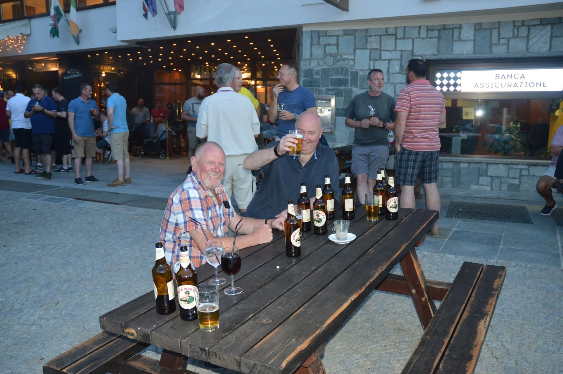 Two men sitting at a picnic table outside, smiling and holding beer glasses, with several beer bottles on the table. Behind them, a group of people are standing and chatting in front of a building with a sign that says 'Banca Assicurazione' and a win