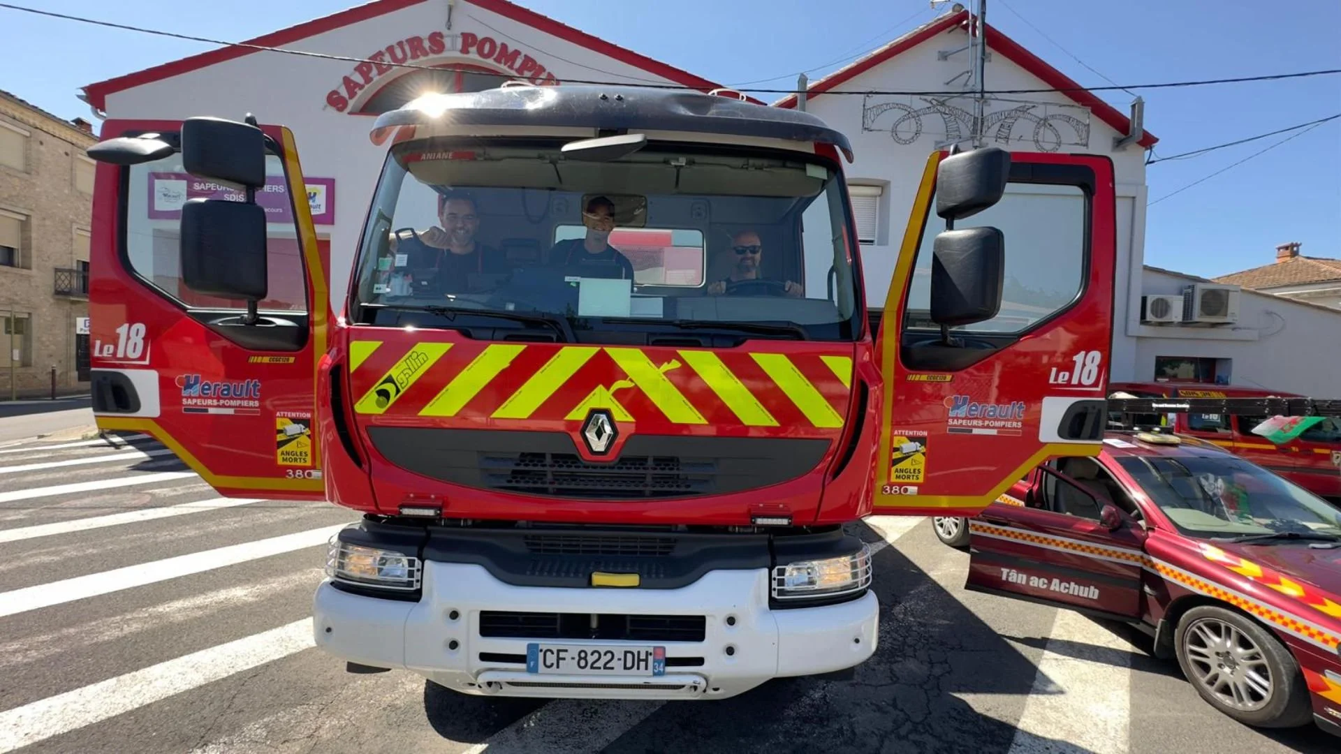 A red French fire truck with yellow reflective markings and a Renault logo, parked with crew inside, in front of a building with the words 'Sapeurs Pompiers.' There is a red car next to it, and a smaller vehicle with similar markings on the right.