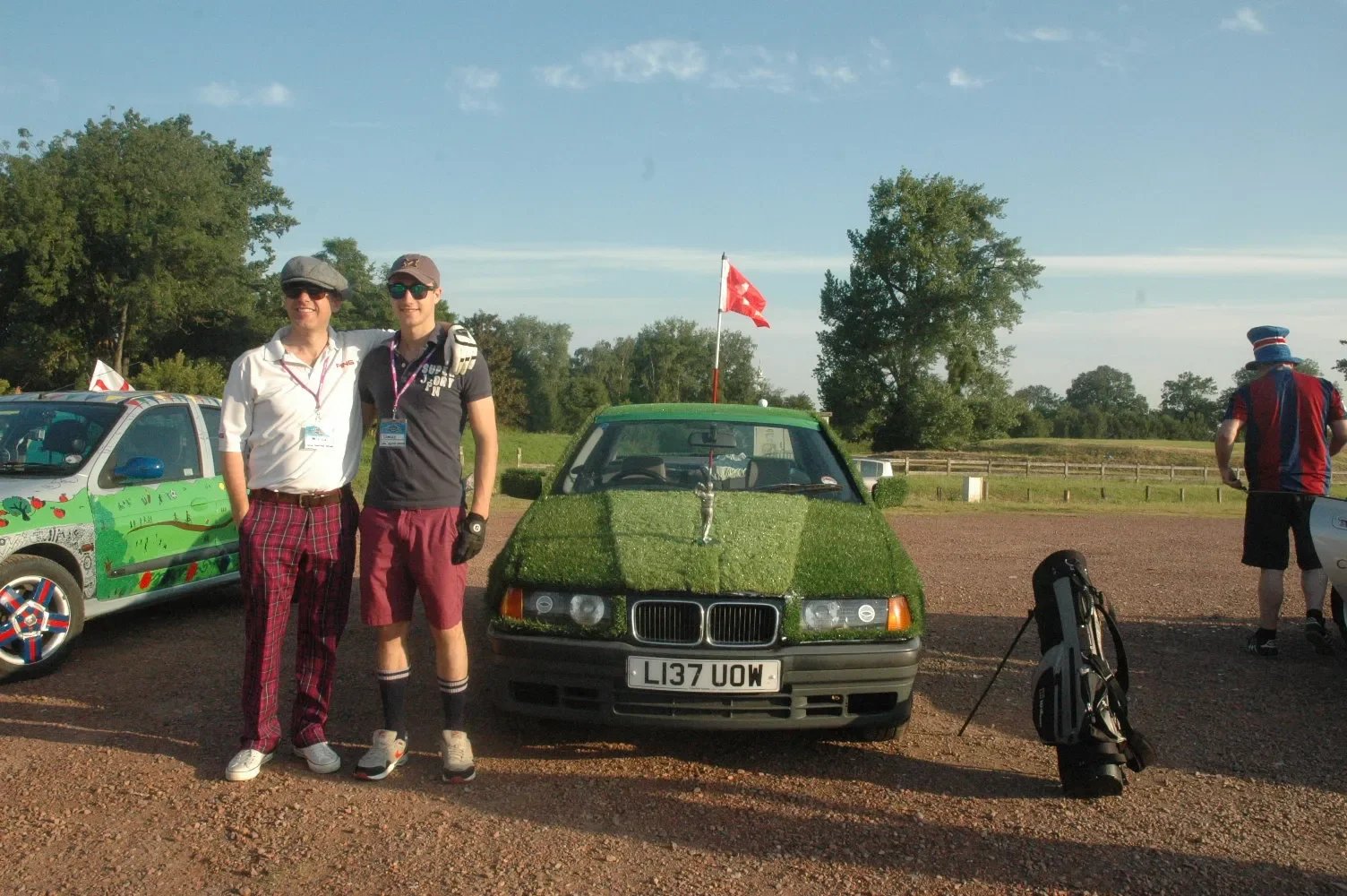 Two men standing outdoors next to a car decorated with grass and a small flag, with other people and cars in the background on a clear day.