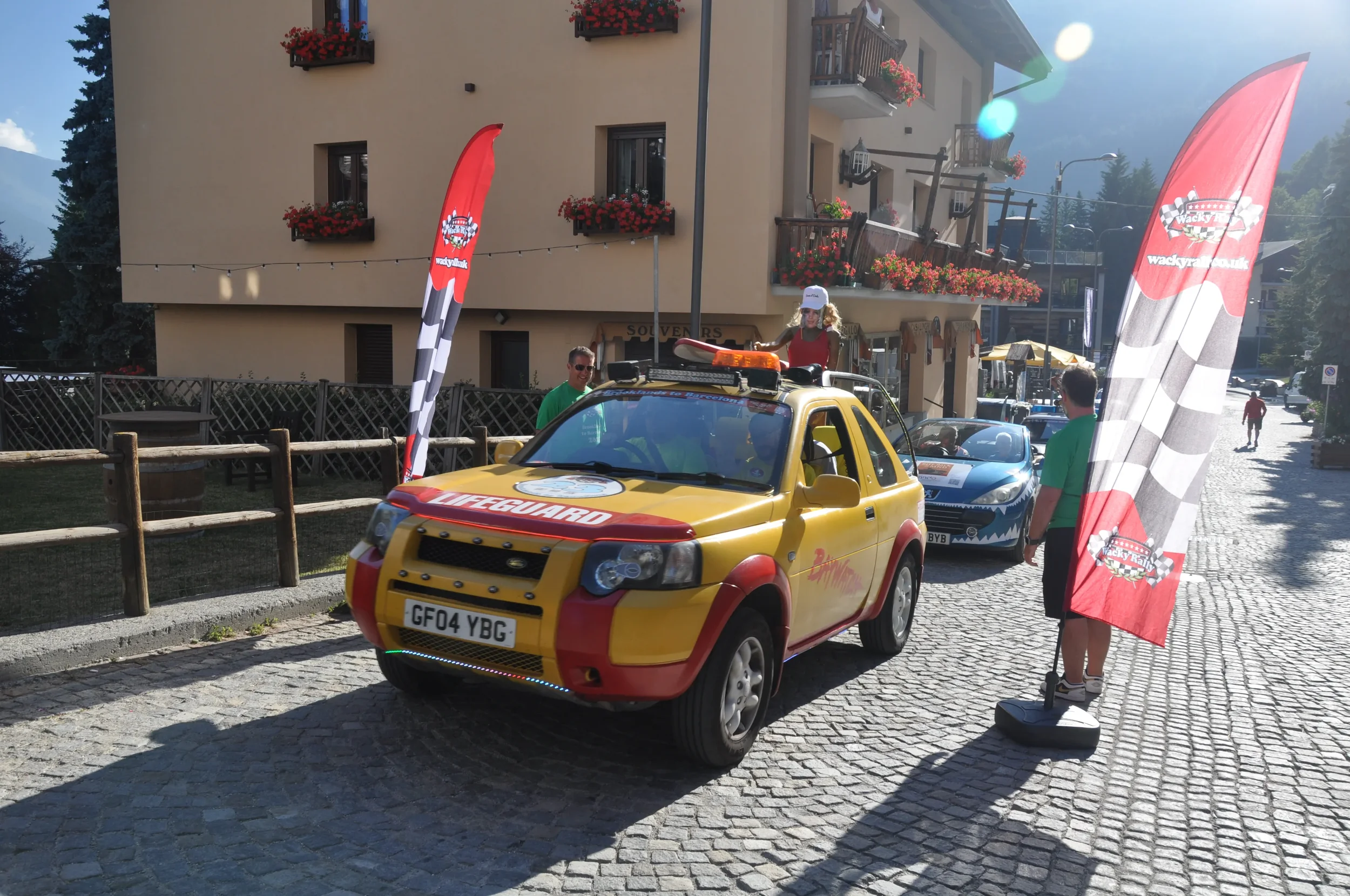 A colorful toy car marked as a lifeguard vehicle is parked on a cobblestone street during an event, with two vertical flags on each side bearing checkered racing pattern and a logo. A woman in a red top and white cap is sitting on top of the toy car,