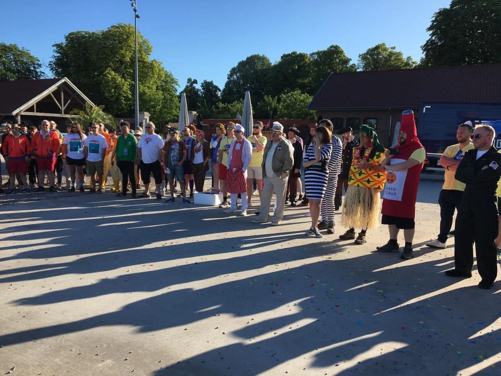Group of people dressed in various costumes gathering outdoors on a sunny day, some wearing striped outfits, a hot dog costume, and other themed attire, standing in a parking lot with long shadows.