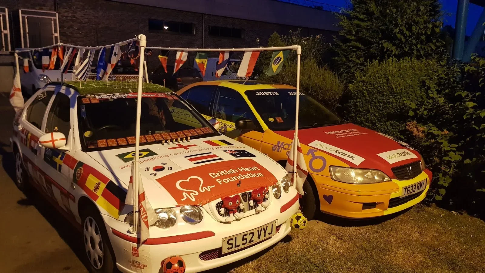 Two race cars decorated with health and charity-themed stickers, flags, and banners, parked next to each other outdoors during the evening. The white car features a prominent British Heart Foundation logo and red cross symbols, while the yellow and r