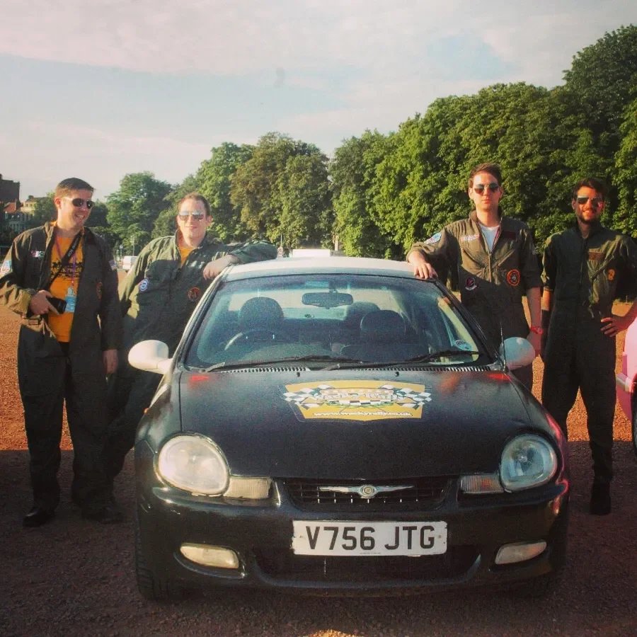 Five young people in racing jackets standing around a black sports car with a police badge decal on the hood, on a dirt field with trees in the background.