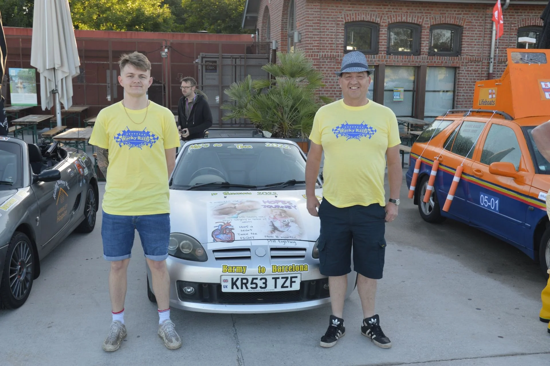Two men wearing yellow T-shirts with a blue checkered flag and the words 'Wacky Rally' embroidered on them, standing in front of a silver car with a banner on the hood that reads 'Barmy to Barcelona'. The car has a license plate 'KR53 TZF'. There are