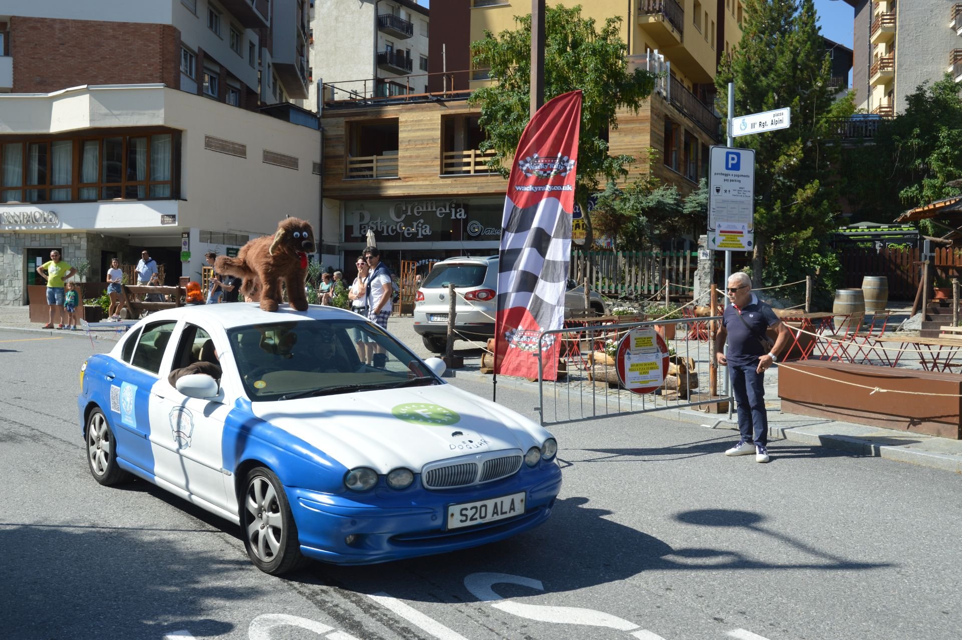 A decorated car with a dog costume on the roof, participating in an outdoor event on a city street. People are gathered around, and a red and black flag stands beside the car.