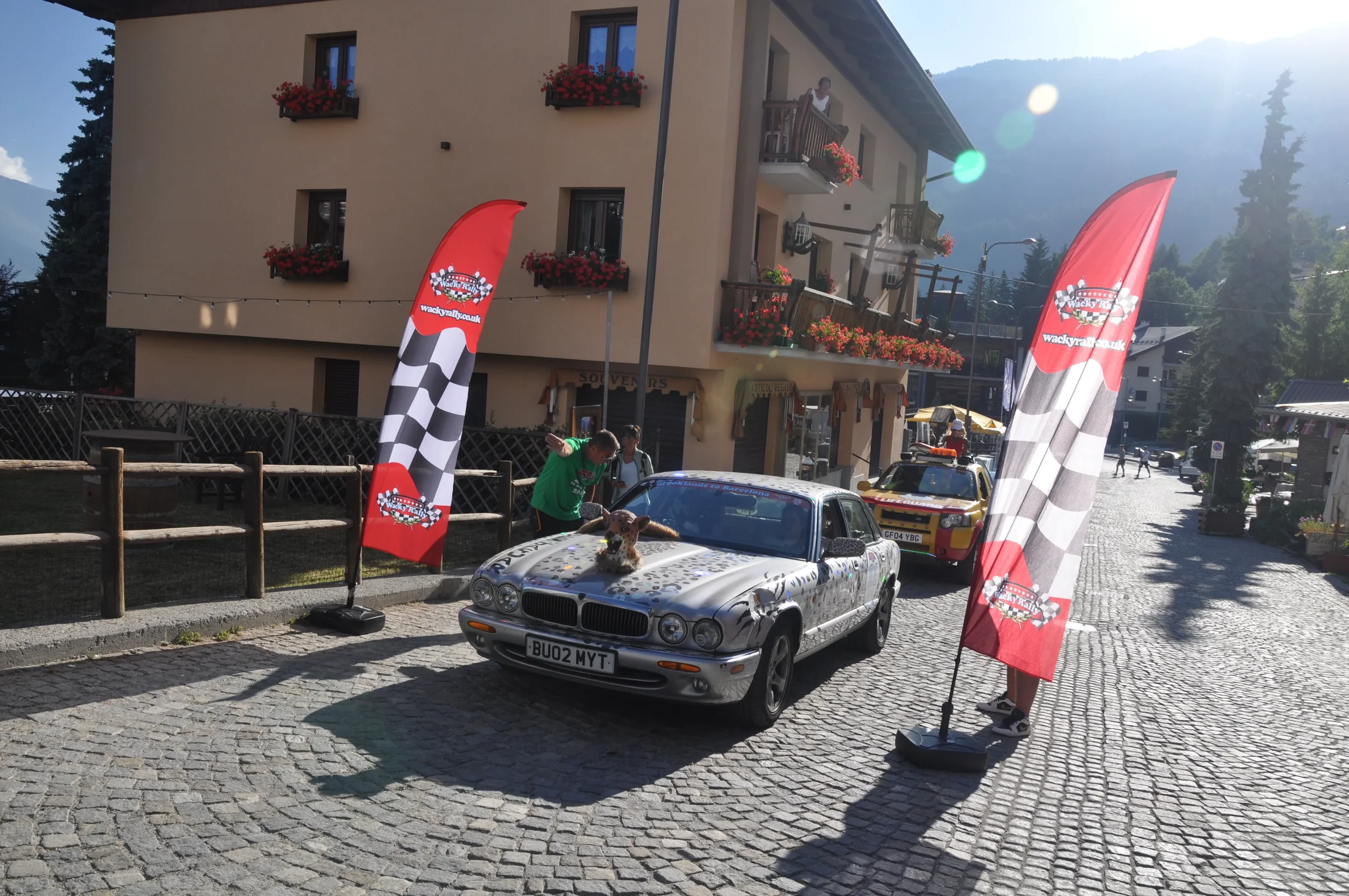 A car decorated with graffiti-style art and a plush animal on its hood, parked between two flags with checkered pattern and a logo, at an outdoor event in a town with cobblestone streets.