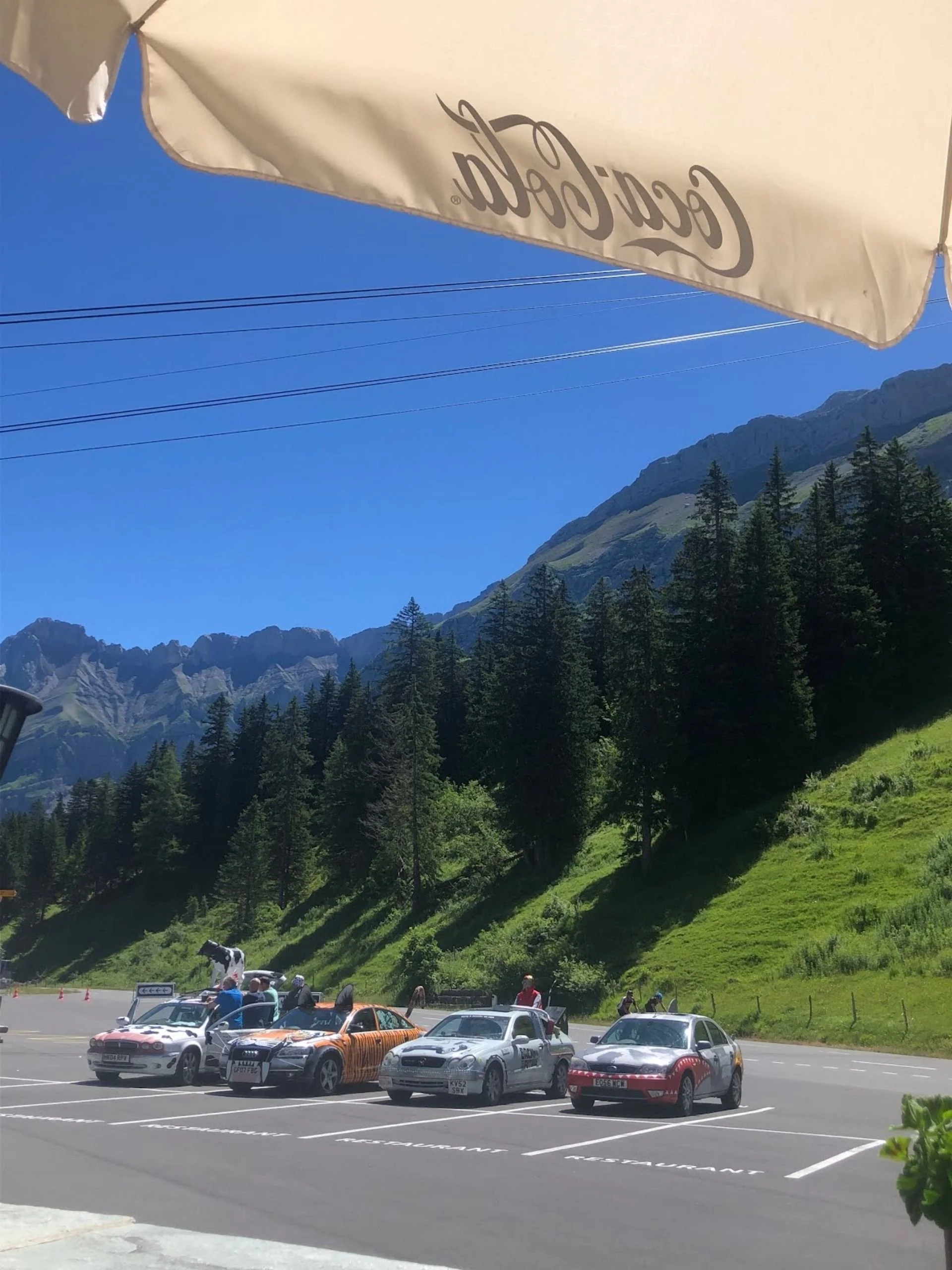 A parking lot with several cars parked, set against a green hillside with tall trees and mountains in the background, under a clear blue sky. An umbrella with the Coca-Cola logo is partially visible at the top of the image.