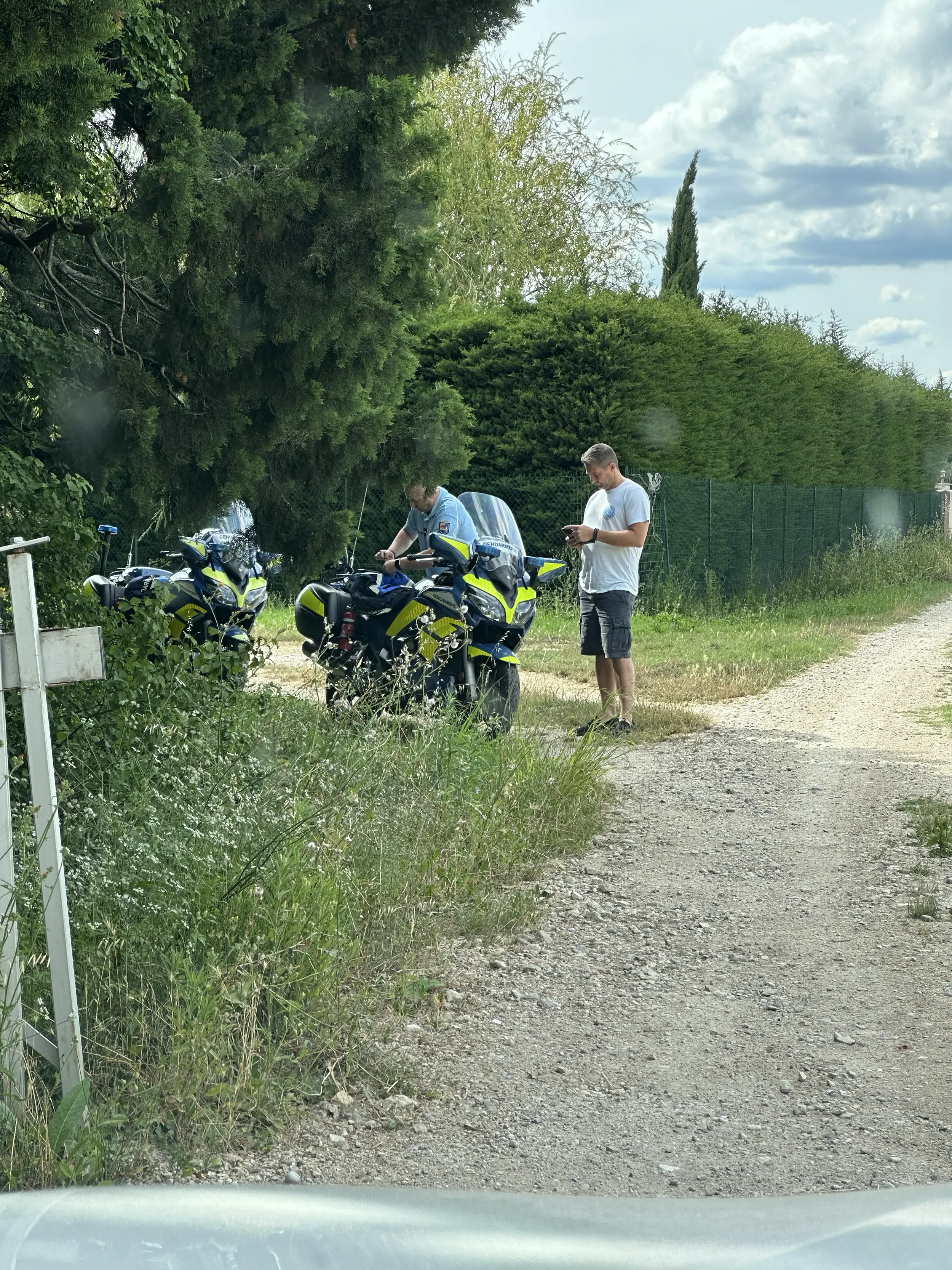 Two police officers and a civilian man are standing next to three police motorcycles on a grassy and dirt roadside with trees and bushes in the background.