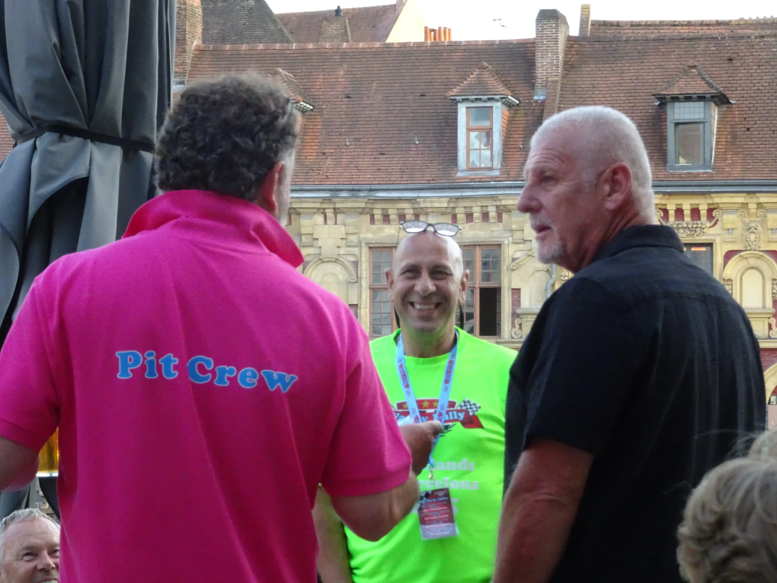Three men are standing outdoors, engaged in conversation; one wearing a pink shirt with 'Pit Crew' on the back, another in a bright yellow shirt with event badges, and the third in a black shirt, with historic buildings in the background.