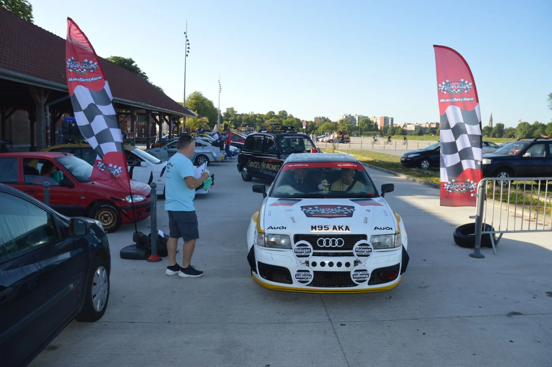 A white rally car with the license plate M95 AKA, parked at an outdoor car event. The car is equipped with additional lights on the front and has racing decals. Two flags with checkered patterns and rally graphics are positioned on either side of the
