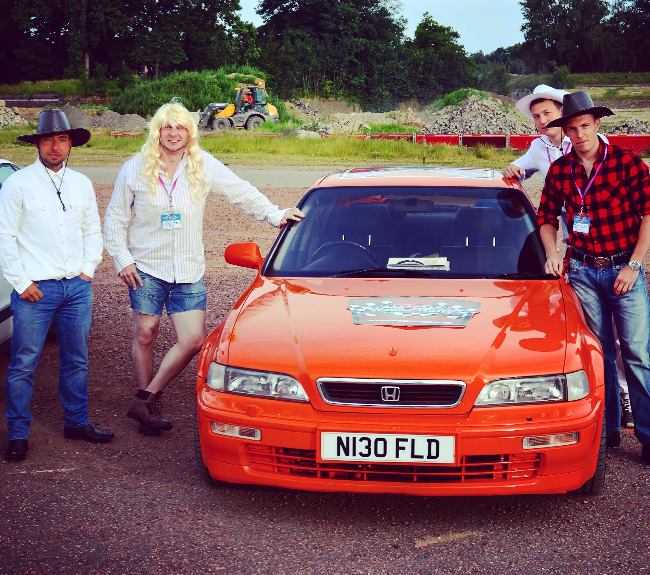 Four men dressed in country-western style clothing and hats stand around a bright orange Honda Civic with license plate N130 FLD, on a gravel lot with construction equipment and trees in the background.