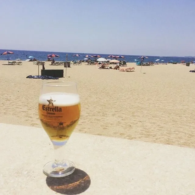 A glass of beer on a table at the beach with sand, umbrellas, and lounge chairs in the background.