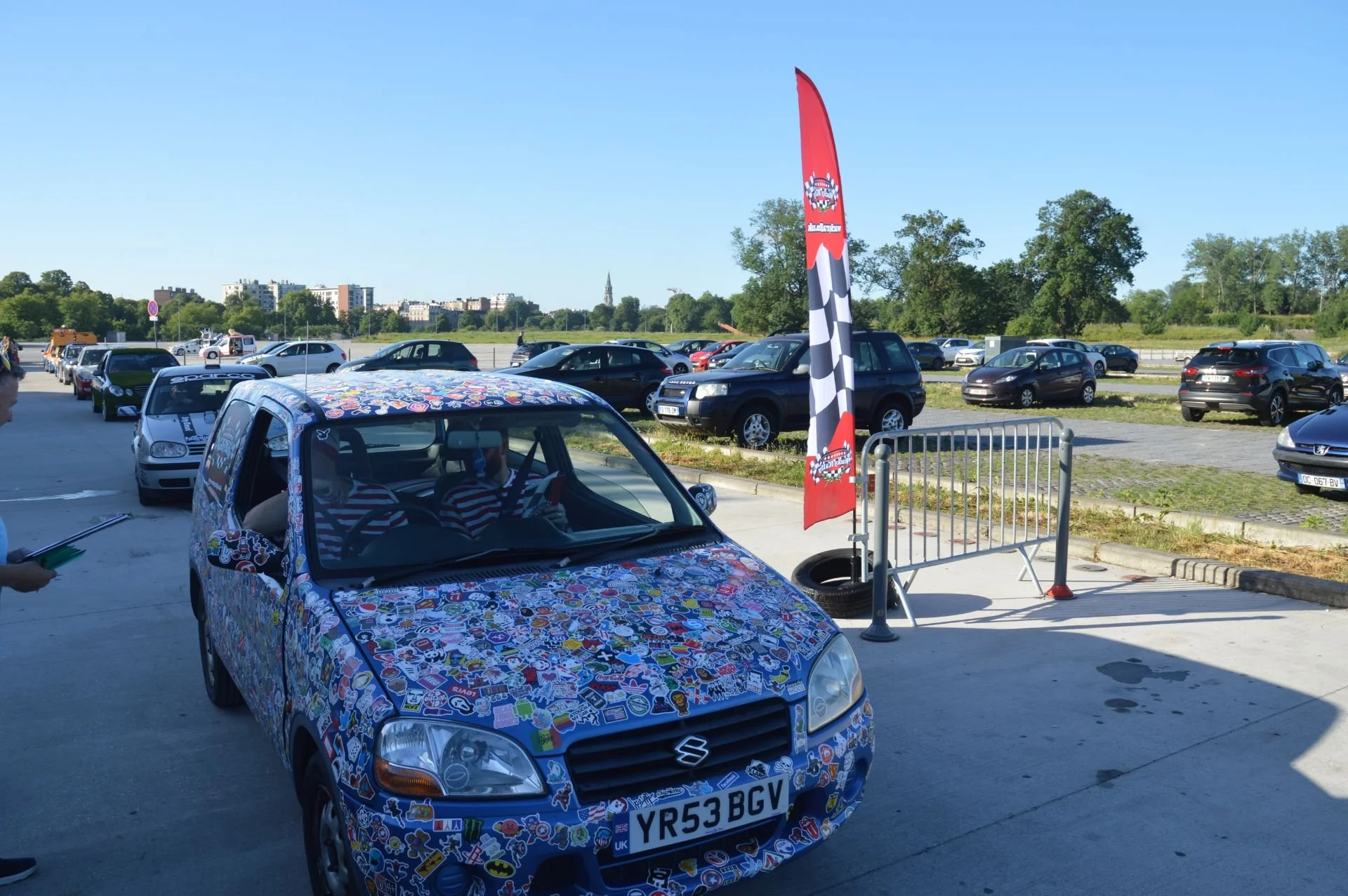 A small blue Suzuki car covered in various colorful stickers, parked in a parking lot at an outdoor event with other cars, a red flag with a checkered pattern, and a metal barrier.