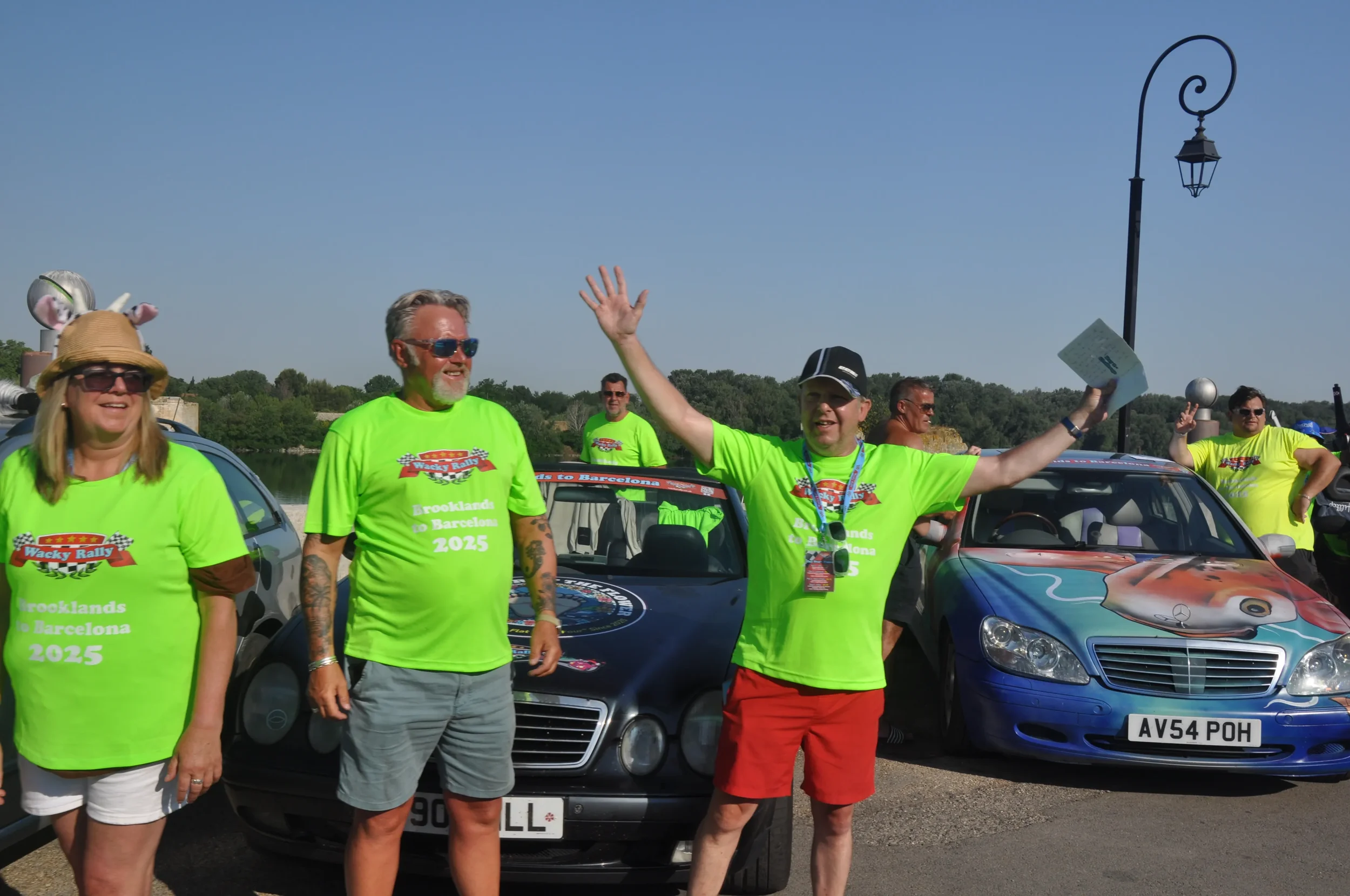 Group of people wearing bright green T-shirts with a rally logo, gathering beside decorated cars at an outdoor event, under clear blue sky.