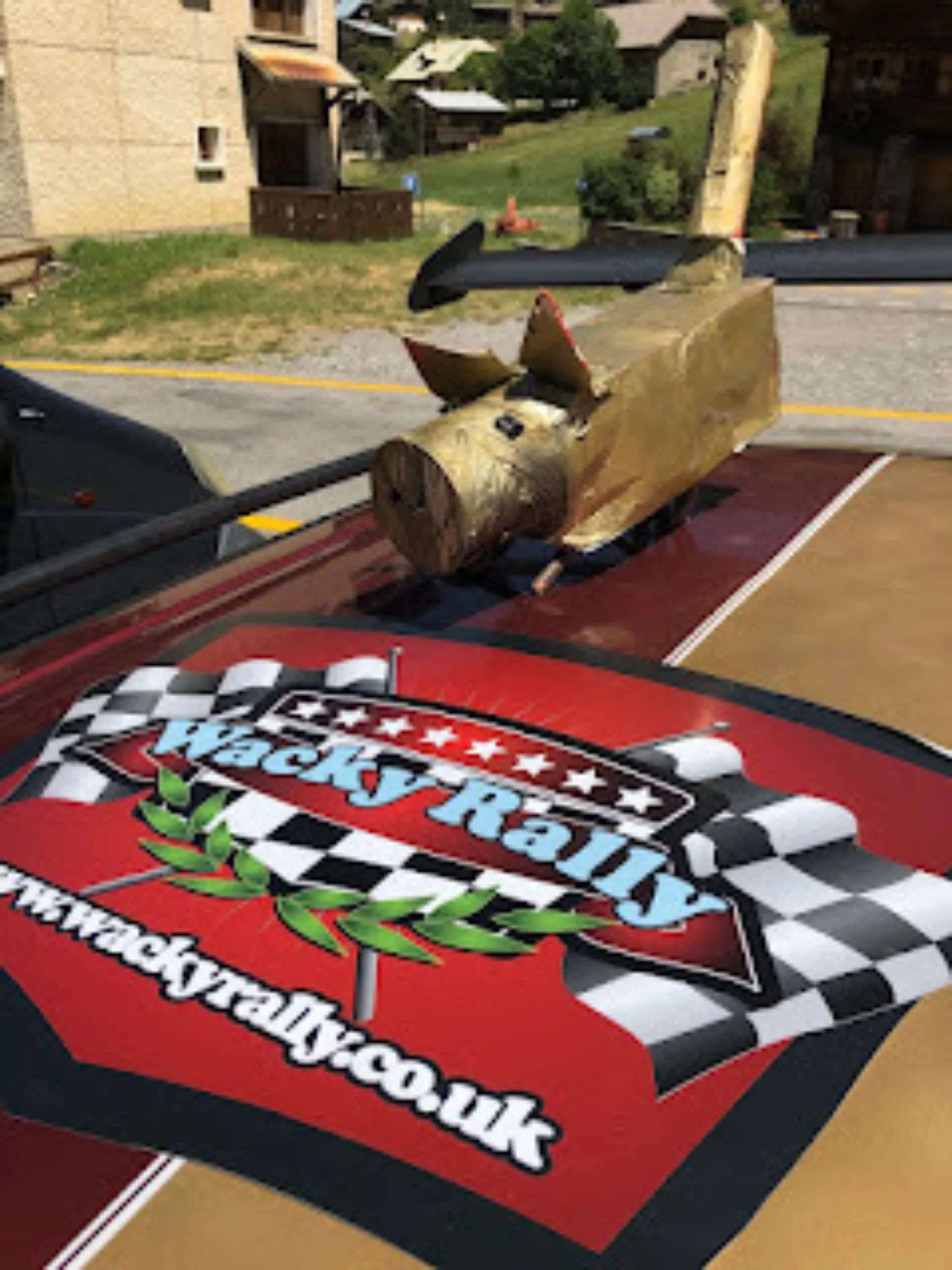 A racing-themed truck with a logo featuring checkered flags and the website 'www.wackyracetyco.uk,' a wooden log with a propeller attached on top, and a competition starting flag in the background.