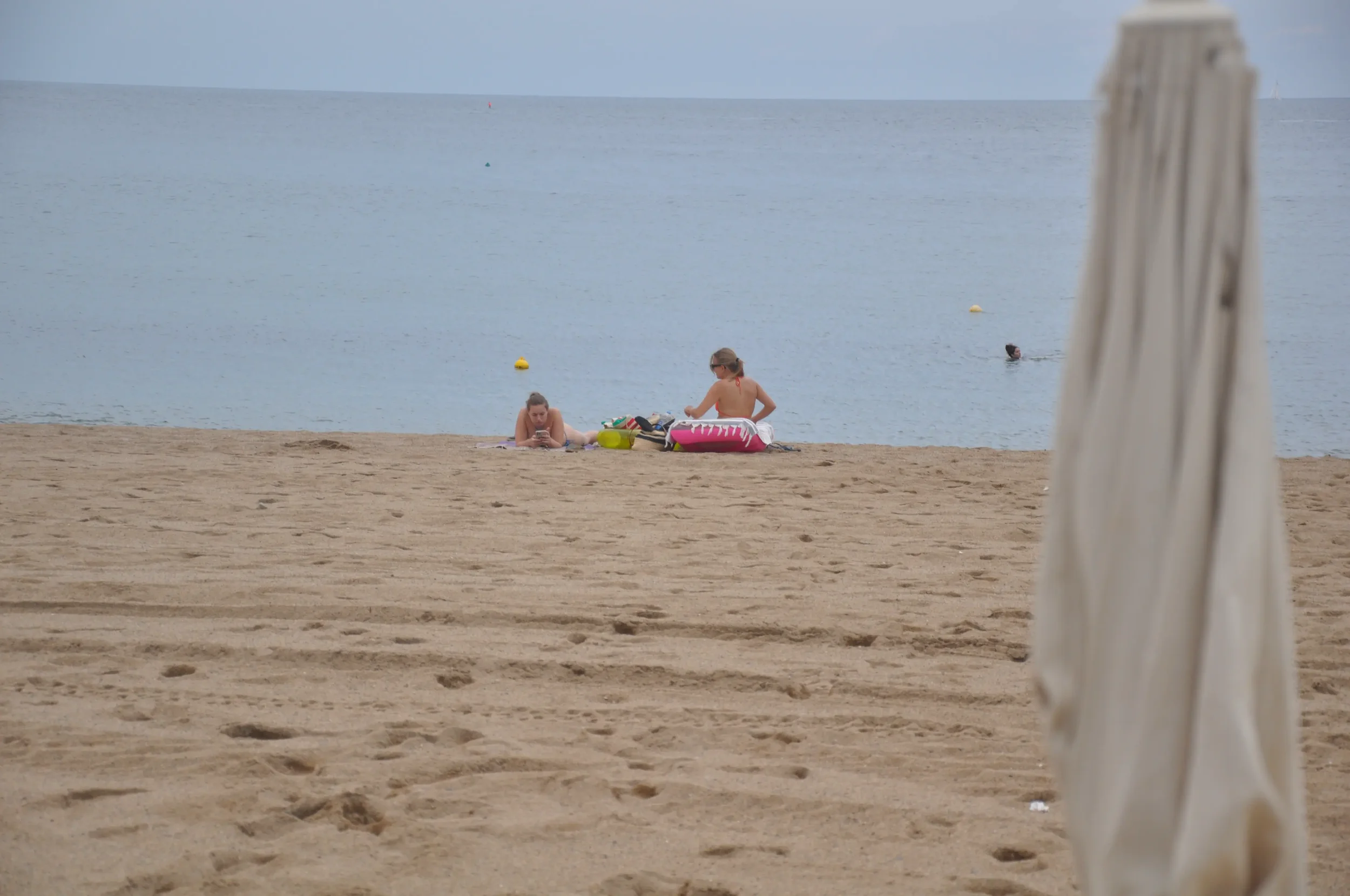 Two women sitting on the sandy beach near the water, with one looking at their phone and the other tending to their belongings, and a person swimming in the ocean in the background, with a partially visible beach umbrella in the foreground.