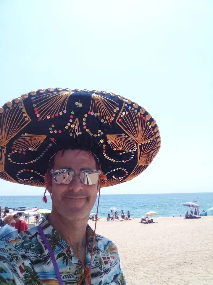 Man wearing sunglasses, a patterned Hawaiian shirt, and a large decorated sombrero taking a selfie on the beach with the ocean, sand, umbrellas, and beachgoers in the background.