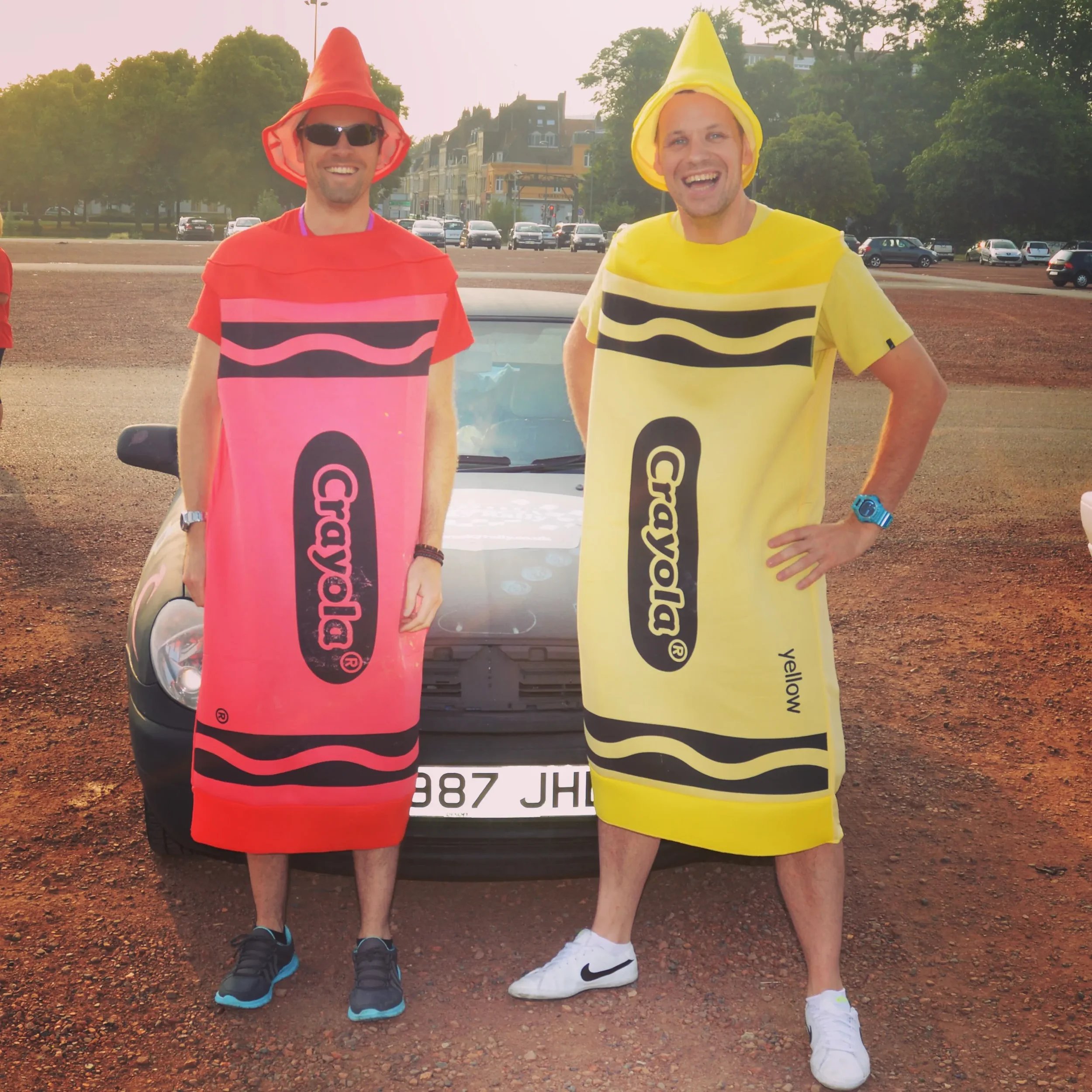 Two men in hotdog costume suits stand in front of a car, smiling. The man on the left wears a pink hotdog costume with the word 'Crayola' on it, sunglasses, and black sneakers. The man on the right wears a yellow hotdog costume with the word 'Crayola