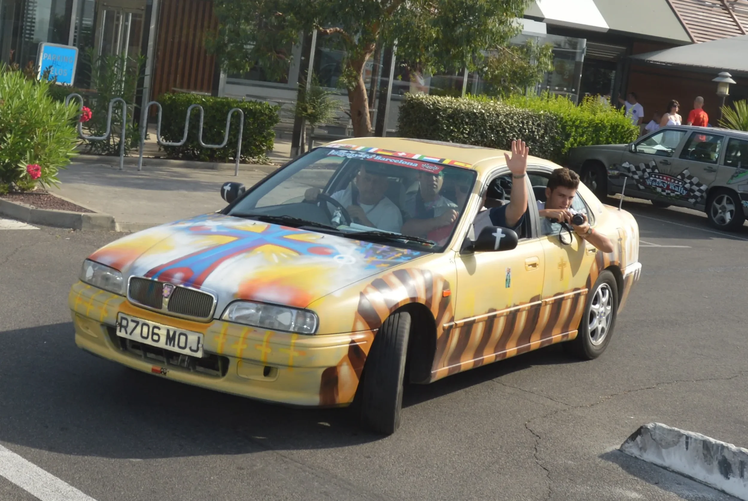 A car painted with religious symbols and colorful design, with a person waving and another taking a photo, parked in a lot near some greenery and buildings.