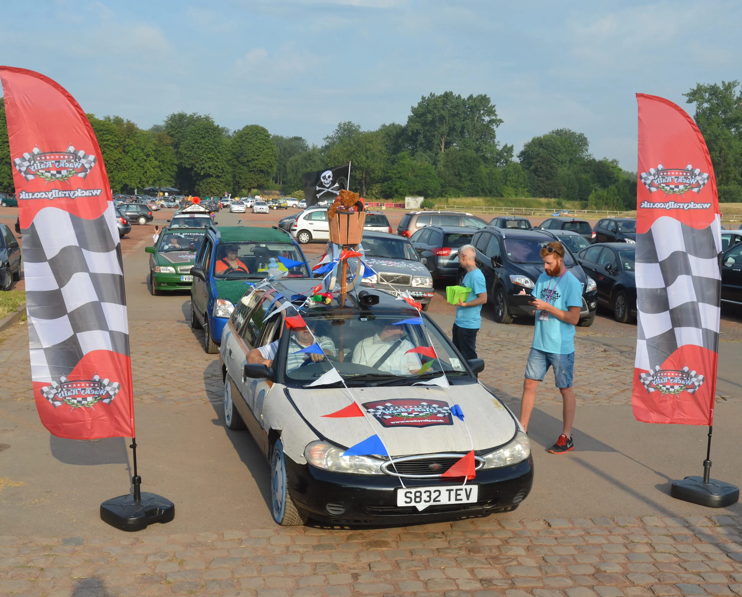 A vintage car decorated with racing flags and banners in a parking lot during a car racing event, with people standing nearby and a large crowd in the background.