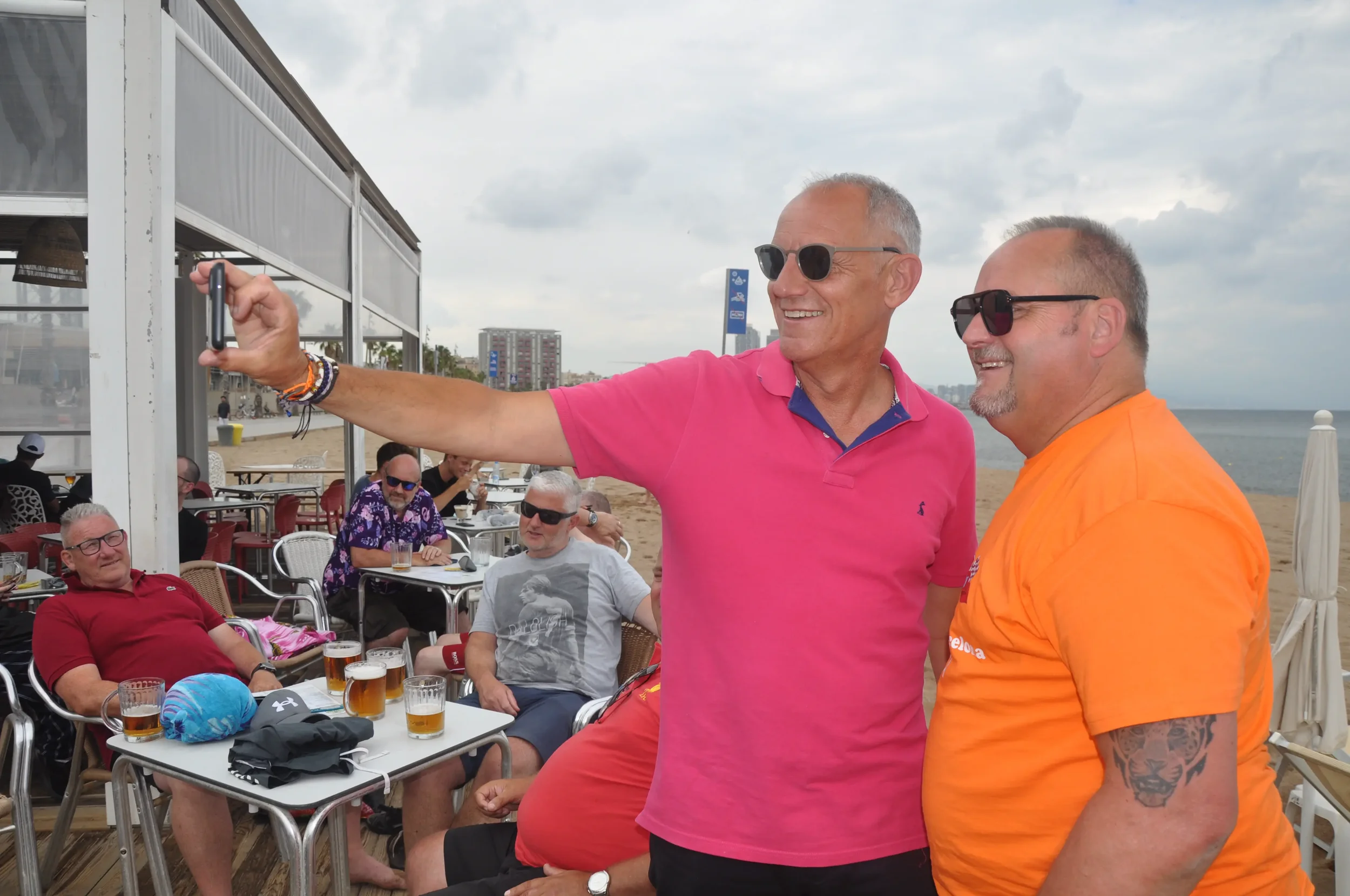 Two smiling men wearing sunglasses taking a selfie on the beach with a group of friends sitting at tables behind them.
