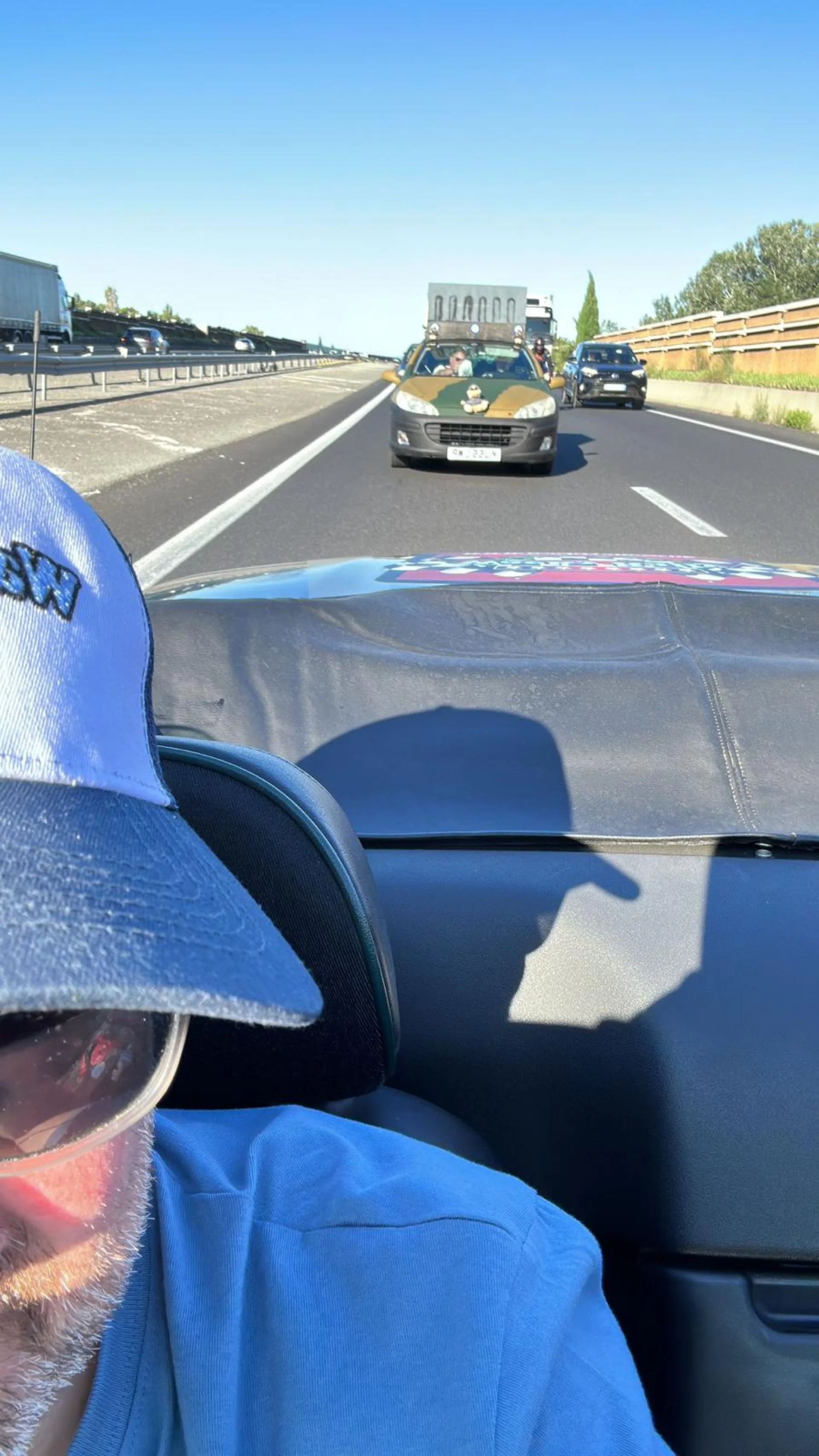 A view from inside a car on a highway, showing the dashboard, a person with a gray beard wearing a blue cap and shirt, and a shadow of a hat on the dashboard. There are other cars on the highway, including one with a camouflage paint job and a large 