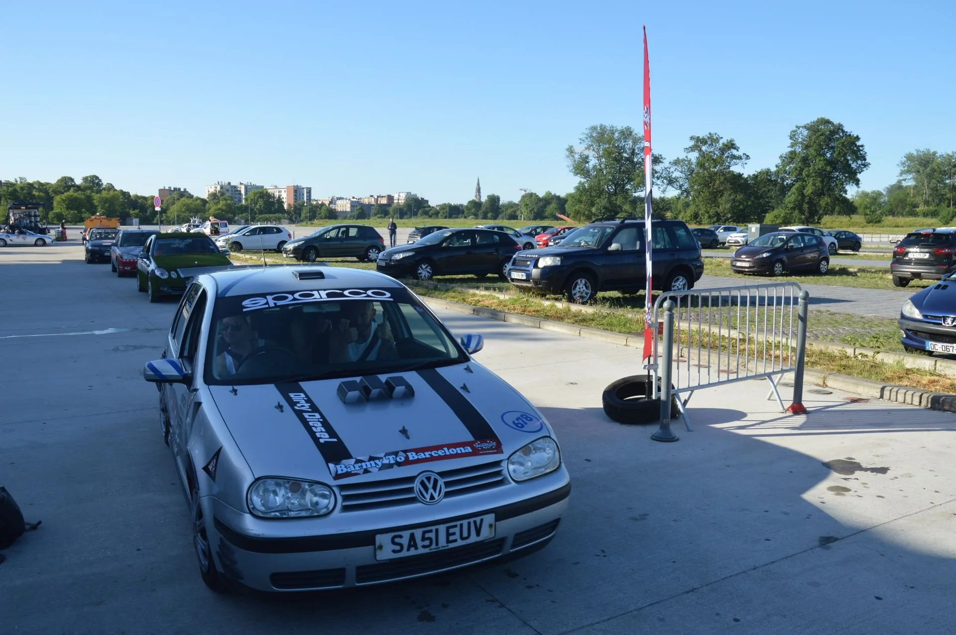 A silver Volkswagen race car with custom decals, parked in a lot with other vehicles under a clear blue sky.